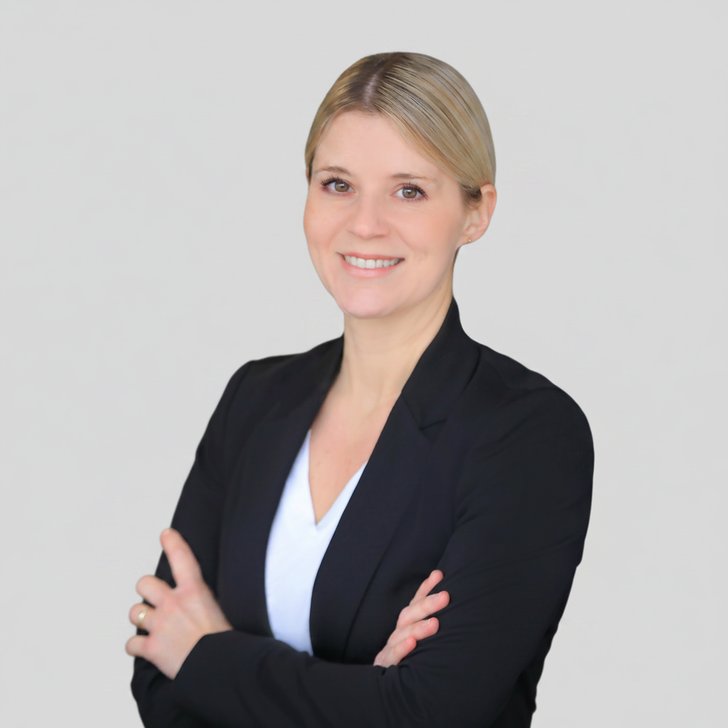 Das Bild zeigt eine Frau mit blondem Haar, die einen schwarzen Blazer und ein weißes Oberteil trägt. Sie hat ihre Arme verschränkt und lächelt. Der Hintergrund ist einfarbig hellgrau. Es handelt sich vermutlich um ein professionelles Porträtfoto, wie es beispielsweise für Bewerbungen oder Unternehmensprofile genutzt wird.