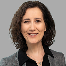 Das Bild zeigt eine Frau mit schulterlangem, braunem Haar, die freundlich lächelt. Sie trägt ein graues Sakko über einer schwarzen Bluse und eine Halskette. Der Hintergrund ist neutral grau und das Bild wirkt wie ein professionelles Porträt.
