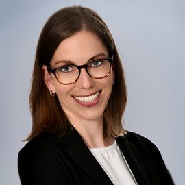 Die abgebildete Person trägt ein helles Oberteil und einen dunklen Blazer. Sie hat mittellanges, glattes Haar und steht vor einem neutralen, hellen Hintergrund.