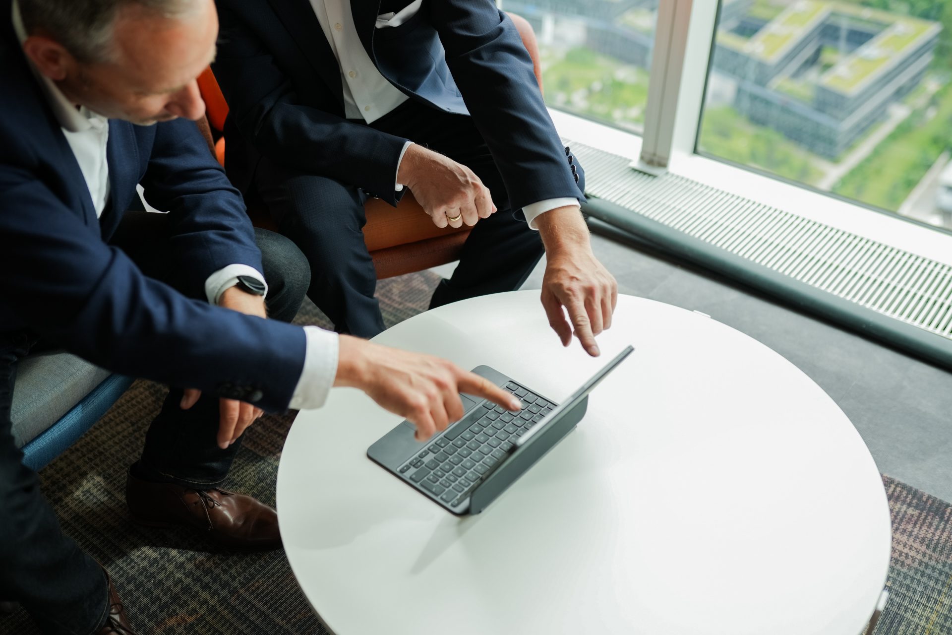 Das Bild zeigt zwei Männer mit Anzug, die an einem runden Tisch sitzen und auf einen Laptop blicken. Einer der Männer zeigt mit seinem Finger auf den Bildschirm des Laptops, während der andere ebenfalls seine Aufmerksamkeit darauf richtet. Die Szene deutet auf eine geschäftliche Besprechung oder Diskussion hin, bei der möglicherweise ein digitales Dokument oder eine Präsentation betrachtet wird. Im Hintergrund ist ein Fenster zu sehen, das einen Blick auf ein modernes Bürogebäude mit begrünten Dachflächen bietet.