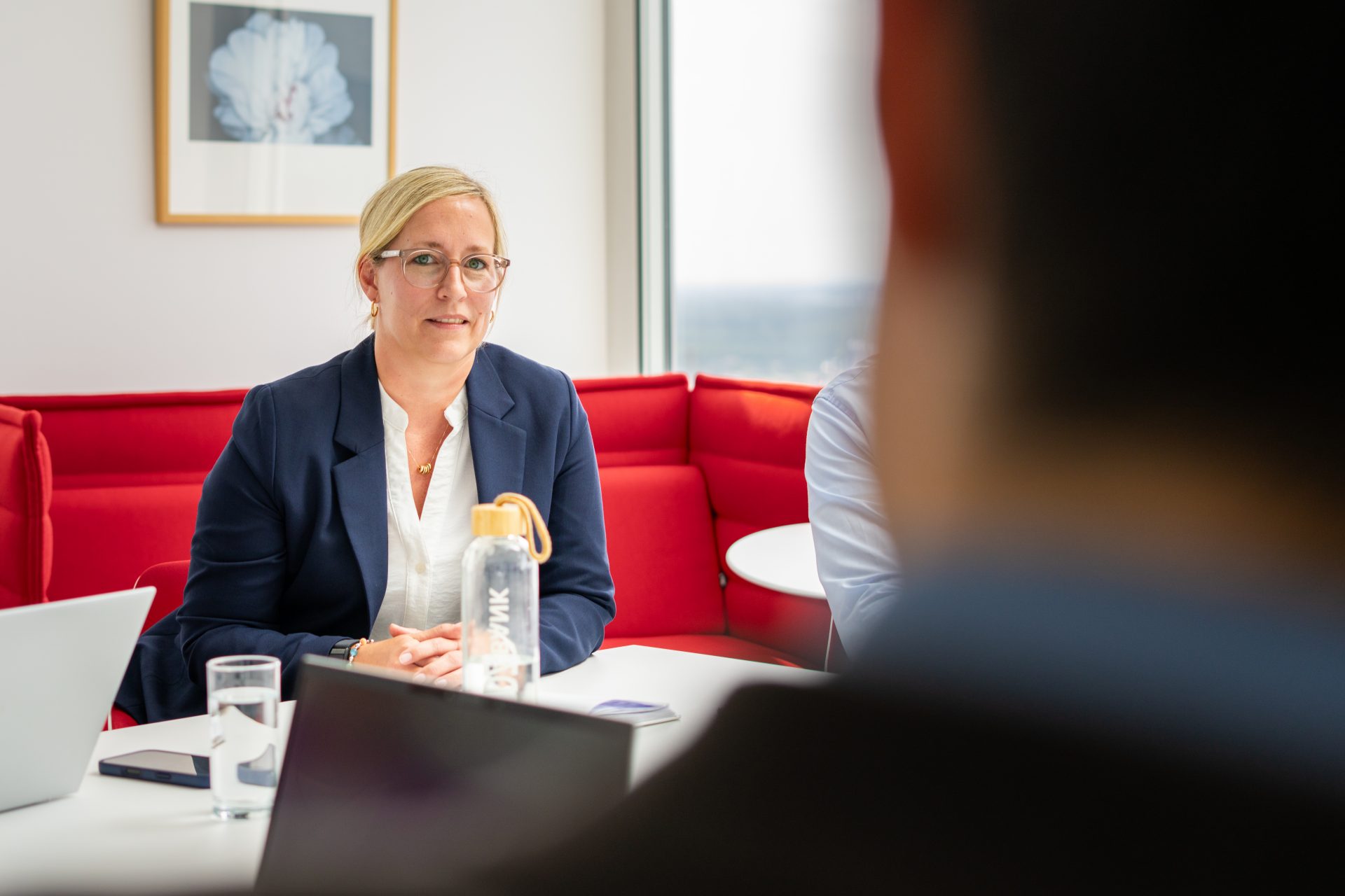 Ein Meeting oder Gespräch in einem modernen Büro oder Arbeitsumfeld, mit mehreren Personen und der Fokus liegt auf einer Person im Vordergrund, die vor einem roten bequemen Sitzbereich sitzt. Auf dem Tisch befinden sich Laptops und ein Getränk, was auf eine Arbeitsbesprechung hindeuten könnte.