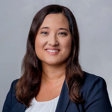 Die Person trägt ein formelles Outfit, bestehend aus einem dunkelblauen Blazer und einer weißen, darunter getragenen Bluse. Der Hintergrund des Bildes ist neutral grau, was oft in professionellen Porträtfotografien verwendet wird.