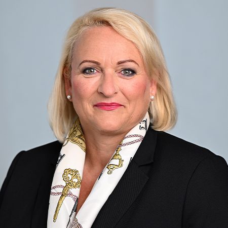 Die Person trägt ein formelles schwarzes Sakko und einen dekorativen Schal mit einem klassischen Muster. Die Ohrringe sind dezent und perlenartig. Der Hintergrund ist neutral, was typischerweise bei professionellen Porträtaufnahmen verwendet wird.