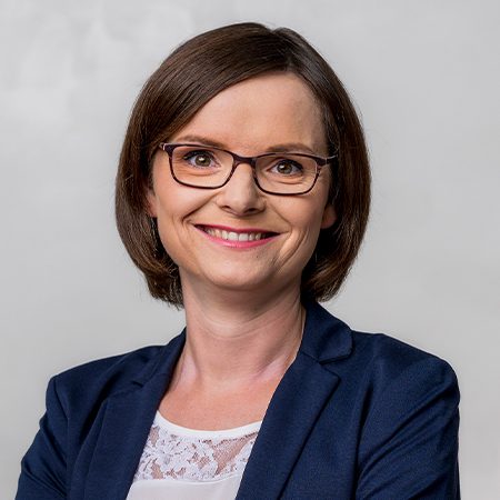 Die Person trägt ein formelles Outfit, bestehend aus einem dunkelblauen Blazer und einer weißen, teilweise durch Spitzenapplikationen verzierten Bluse.