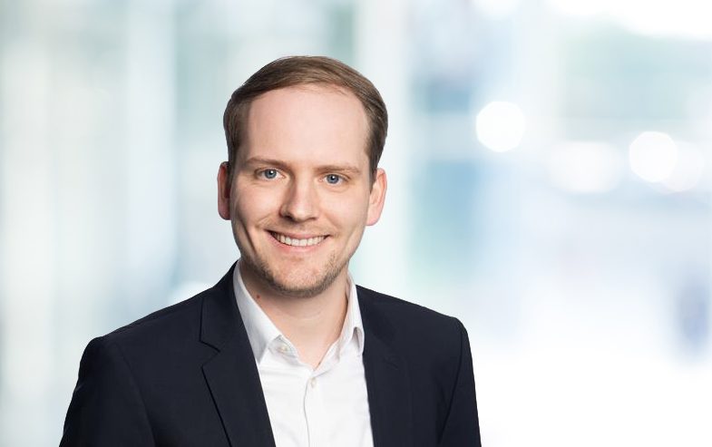 Ein Mann in einem formellen Anzug vor einem verschwommenen Hintergrund.