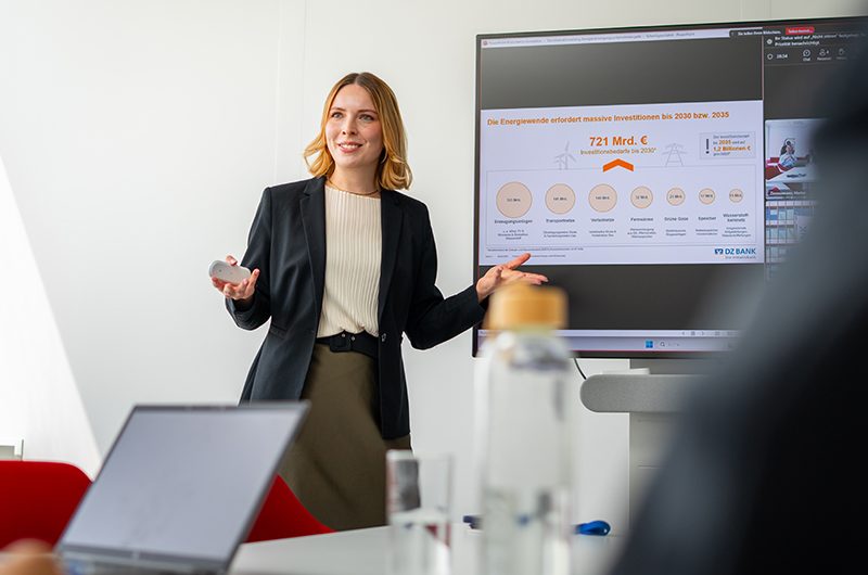 Das Bild zeigt eine Präsentation in einem Büro- oder Meetingraum. Eine Person steht vor einem Bildschirm, auf dem eine Folie mit einem Diagramm und Zahlen bezüglich "ES6 Compliance Tech Mitigation Identification 2024" zu sehen ist. Auf dem Tisch liegen Laptops und eine Wasserflasche.