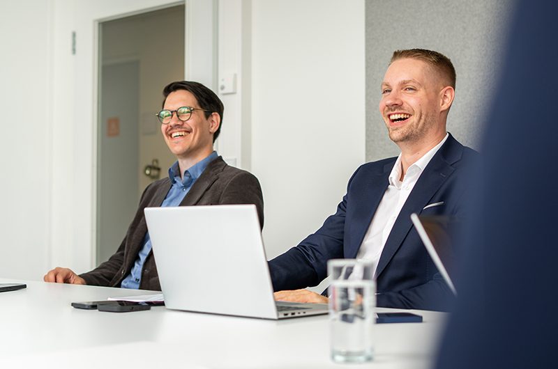 Das Bild zeigt ein formelles Geschäftstreffen oder ein Meeting, bei dem mehrere Personen an einem Tisch sitzen. Es gibt einen Laptop, ein Glas Wasser und Schreibutensilien, die darauf hinweisen, dass es sich um eine berufliche oder geschäftliche Umgebung handelt.