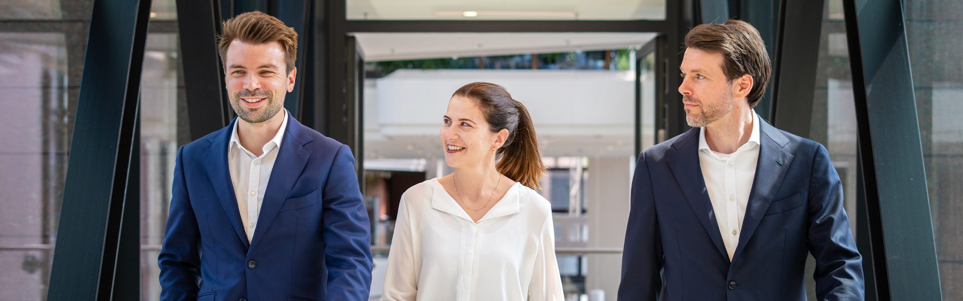 Drei Personen in formeller Kleidung, die einen Flur oder einen Übergang entlang gehen.