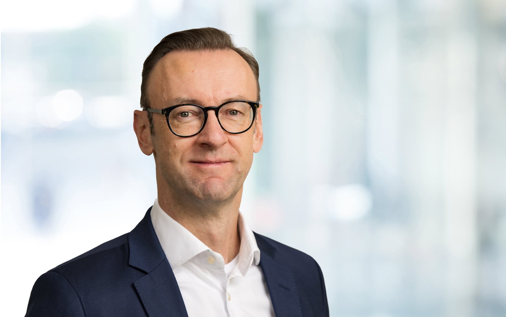 Das Bild zeigt einen Mann mit Brille in einem Anzug und weißem Hemd. Der Hintergrund ist unscharf und wirkt wie eine helle, moderne Innenraumatmosphäre.