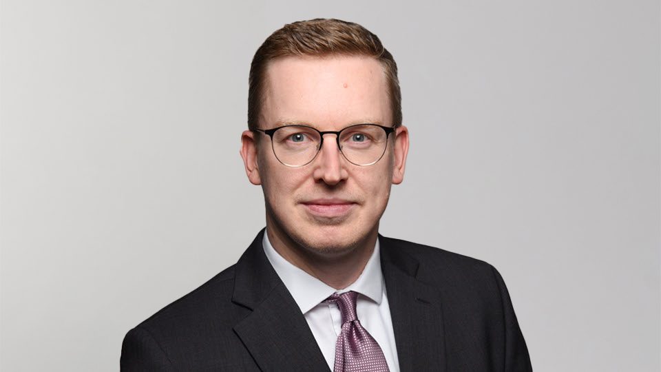 Die abgebildete Person trägt einen dunklen Anzug mit einem weißen Hemd und einem rötlich gemusterten Schlips.