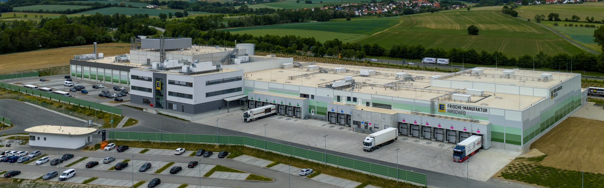 Das Bild zeigt ein modernes Gebäude, das offenbar ein Lagerhaus oder eine Logistikzentrale darstellt. Auf dem Gelände befinden sich mehrere Lastwagen mit Anhängern, die scheinbar auf das Beladen oder Entladen warten. Das Gebäude ist von Parkplätzen umgeben, die für Mitarbeiter oder Besucher des Standorts gedacht sein könnten.
**Hinweise im Bild:**
1. **Architektur:** Die Bauweise des Gebäudes deutet auf eine industrielle oder kommerzielle Nutzung hin.
2. **Beschilderung:** Es gibt sichtbare Hinweise auf ein Unternehmen oder eine Marke am Gebäude.
3. **Umgebung:** Das Bild zeigt ländliches Gebiet im Hintergrund, was auf eine Lage außerhalb urbaner Zentren hinweisen könnte.
4. **Infrastruktur:** Die Organisation der Straße, Parkplätze und Laderampen ist typisch für Einrichtungen, die für große logistische Operationen konzipiert sind.
**Funktion:**
- **Lagerung:** Da Lagerhäuser typischerweise für die Speicherung und Distribution von Waren verwendet werden, ist es wahrscheinlich, dass diese Einrichtung eine ähnliche Funktion hat.
- **Transportlogistik:** Der Fokus auf LKWs, Ladebereiche und Abtransport deuten auf eine logistische Drehscheibe hin.
Zusammenfassend lässt sich sagen, dass das Bild eine logistische oder industrielle Einrichtung zeigt, die in der Lagerung und Verwaltung von Waren tätig ist, möglicherweise mit einem Fokus auf Großhandel oder Massenvertrieb. Das Umfeld und die baulichen Merkmale unterstützen diese Interpretation.