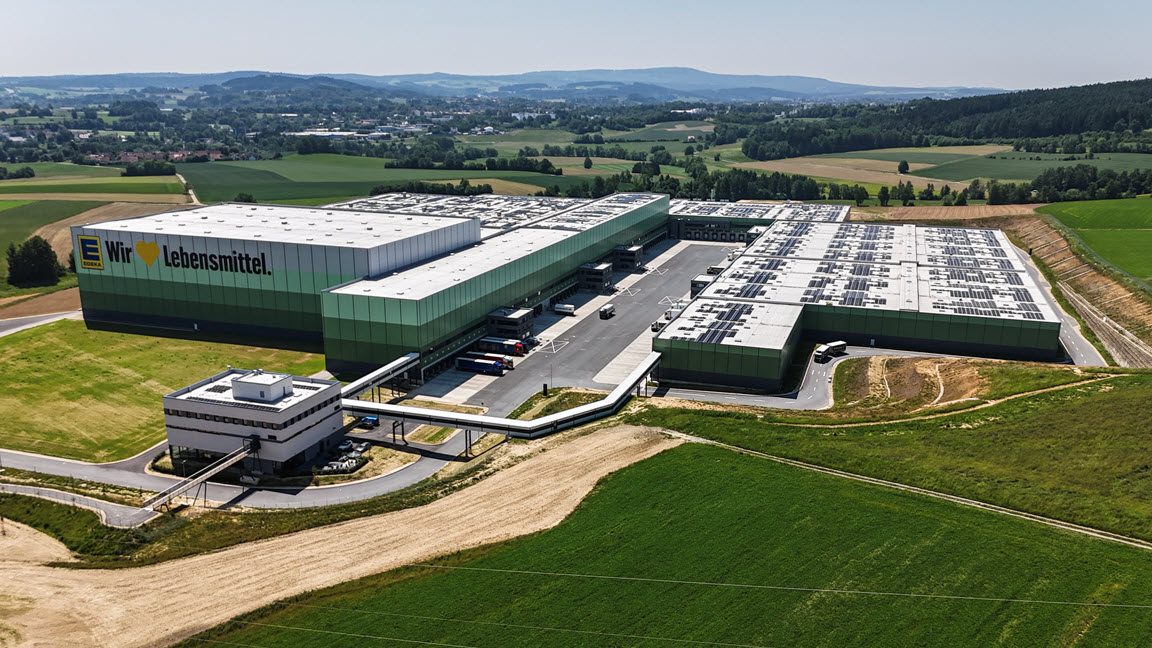 Das Bild zeigt ein großes Gebäude, das ein modernes Lagerhaus oder Distributionszentrum darstellt. Es ist in einer ländlichen Umgebung mit grünen Feldern und Hügeln im Hintergrund eingebettet. Auf dem Gebäude ist das Logo der Firma "C&A" zu sehen, die für Bekleidung bekannt ist, zusammen mit dem Schriftzug "logistics center". Dies deutet darauf hin, dass es sich um ein Logistikzentrum handelt, das für die Lagerung und Distribution von Waren, vermutlich Textilien, genutzt wird. Die Architektur des Gebäudes ist zweckmäßig mit einem klar strukturierten Design und weitläufige Anlagen scheinen gut organisiert für den logistischen Betrieb zu sein.