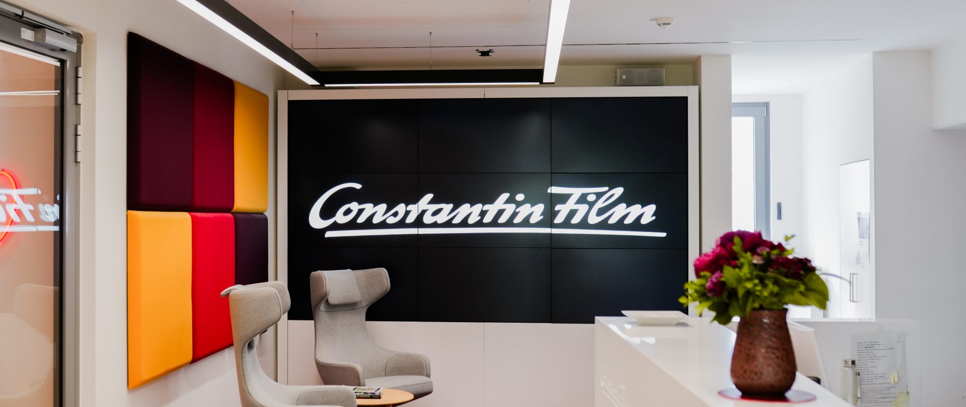 Das Bild zeigt den Empfangsbereich eines Bürogebäudes oder Unternehmens, das mit Constantin Film verbunden ist. Constantin Film ist eine bekannte Filmproduktionsgesellschaft, die für die Herstellung und den Vertrieb von Filmen verantwortlich ist. Der Bereich ist modern gestaltet und enthält einen Bildschirm mit dem Namen der Firma, sowie stilvolle Möbelstücke und dekorative Blumen.