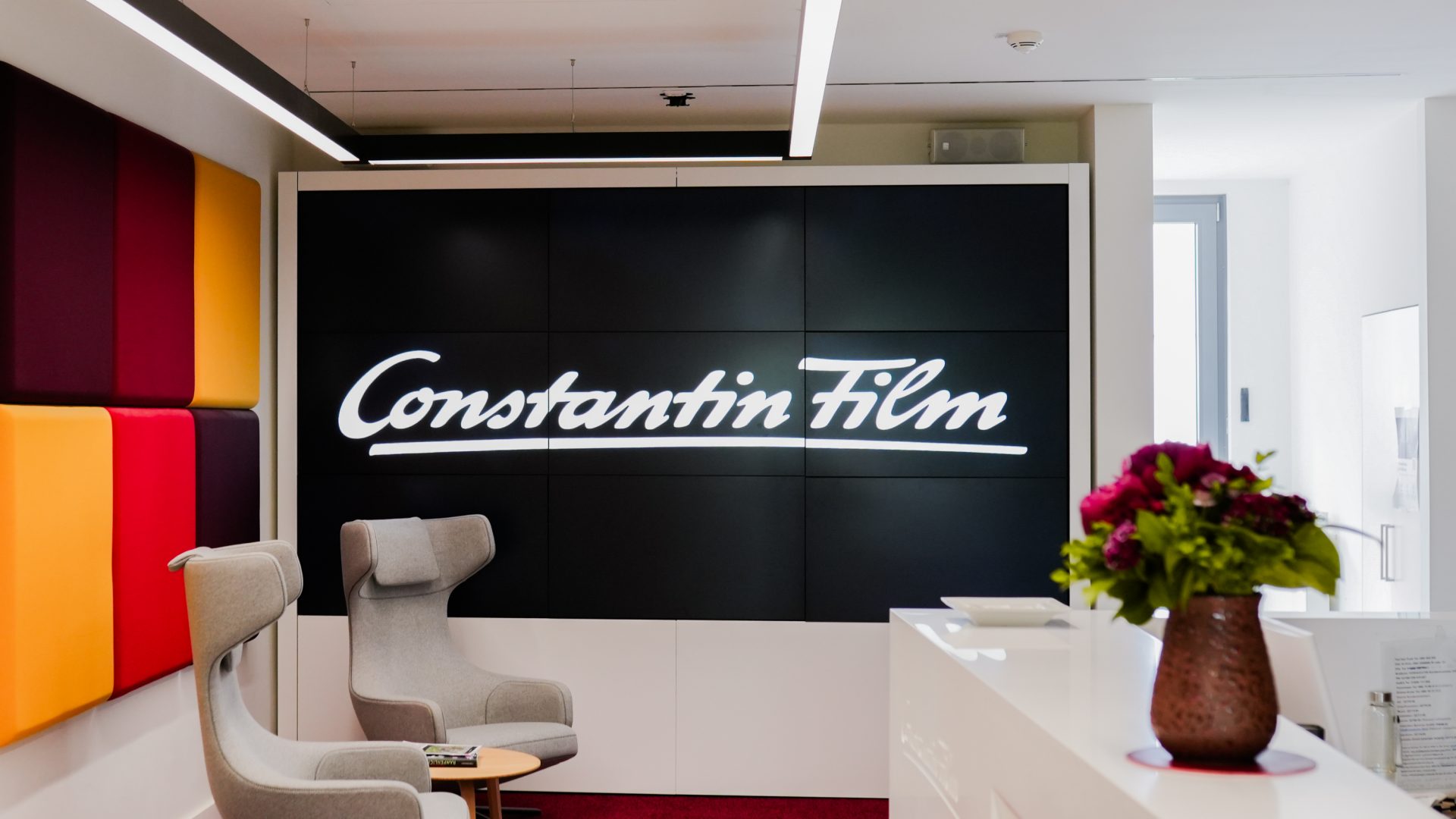 Das Bild zeigt den Empfangsbereich eines Bürogebäudes oder Unternehmens, das mit Constantin Film verbunden ist. Constantin Film ist eine bekannte Filmproduktionsgesellschaft, die für die Herstellung und den Vertrieb von Filmen verantwortlich ist. Der Bereich ist modern gestaltet und enthält bequeme Sitzgelegenheiten, dekorative Wandpaneele und einen Blumenstrauß auf einem Tisch. Der Schriftzug "Constantin Film" auf einem großen Bildschirm oder Panel stellt das Hauptmerkmal des Bildes dar und verweist direkt auf die Identität des Unternehmens.