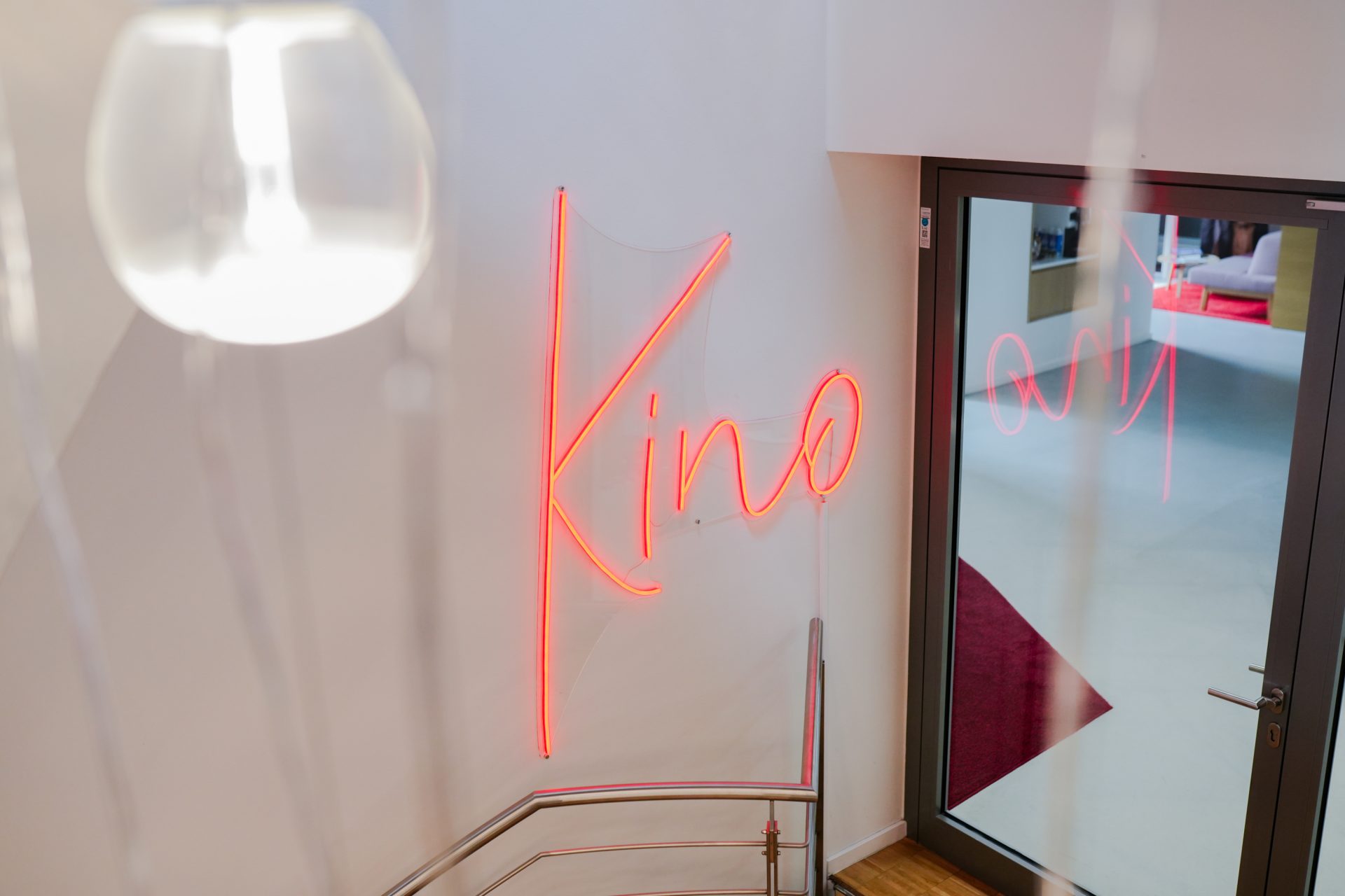 Das Bild zeigt den Eingang oder einen Bereich eines Kinos. Dies wird durch das Wort "Kino", das in roter Neonbeleuchtung geschrieben ist, bestätigt. Die hellen Neonfarben heben sich von der schlichten weißen Wand dahinter ab und verleihen dem Raum ein modernes und einladendes Ambiente. Rechts ist eine Glastür zu sehen, die möglicherweise zu einem Kinosaal oder einem anderen Bereich des Kinos führt. Der Raum scheint sauber und gepflegt zu sein, mit einem Fokus auf einfache, stilvolle Dekorationen, die ein angenehmes Erlebnis für Kinobesucher bieten.