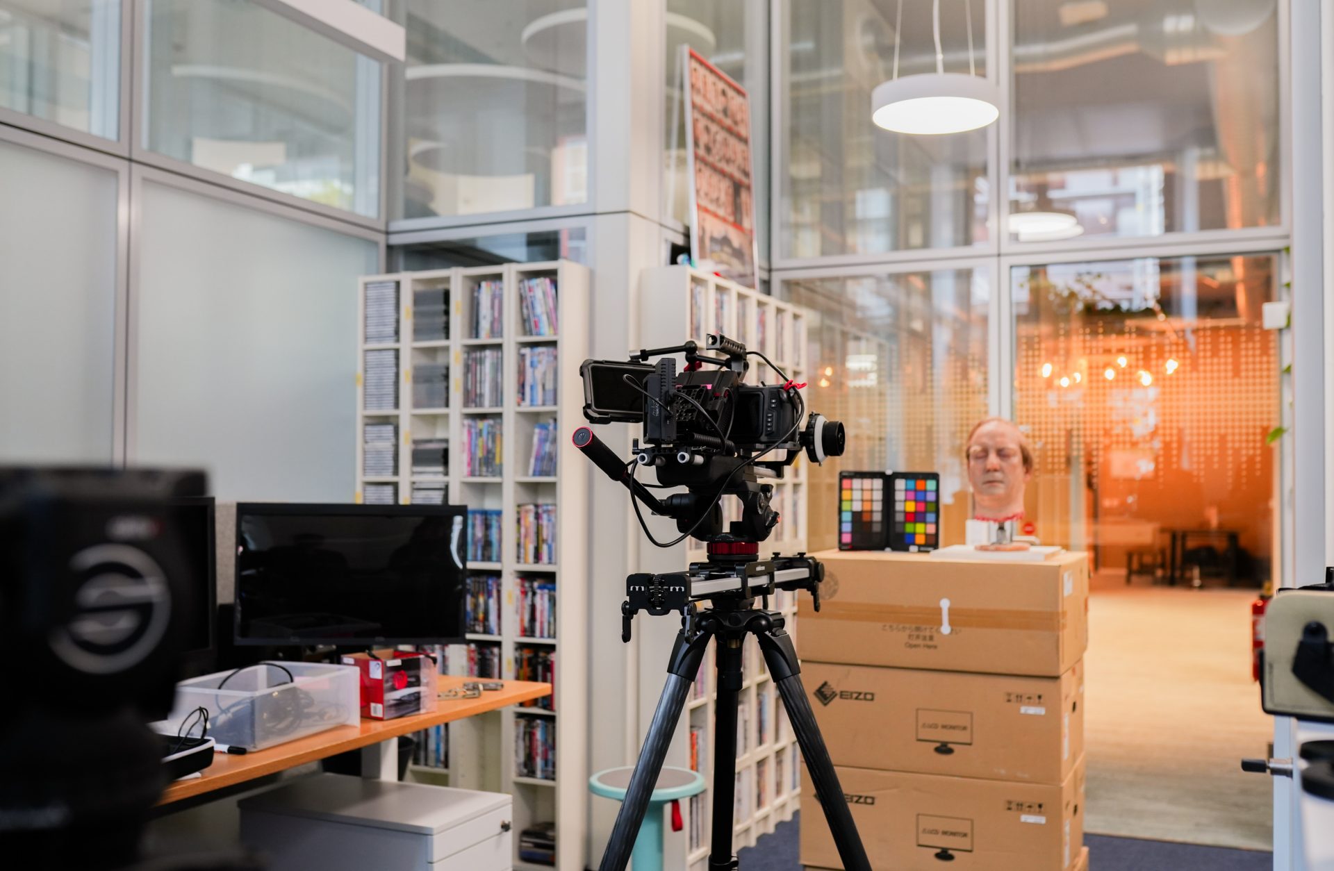 Das Bild zeigt ein Fotostudio oder einen professionellen Büroraum, der für die Medienproduktion eingerichtet ist. Im Vordergrund sieht man eine Kamera auf einem Stativ, scheinbar bereit für eine Aufnahme. Im Hintergrund ist eine Person zu erkennen, die vor einem Stapel von Kartons oder Boxen steht, möglicherweise als Modell oder für ein Testshooting. Der Raum hat eine moderne Einrichtung mit Regalen befüllt mit DVDs oder Bücher und eine große Fensterfront, die für Tageslicht sorgt. Lichter hängen von der Decke und der Raum vermittelt eine kreative und professionelle Atmosphäre.
