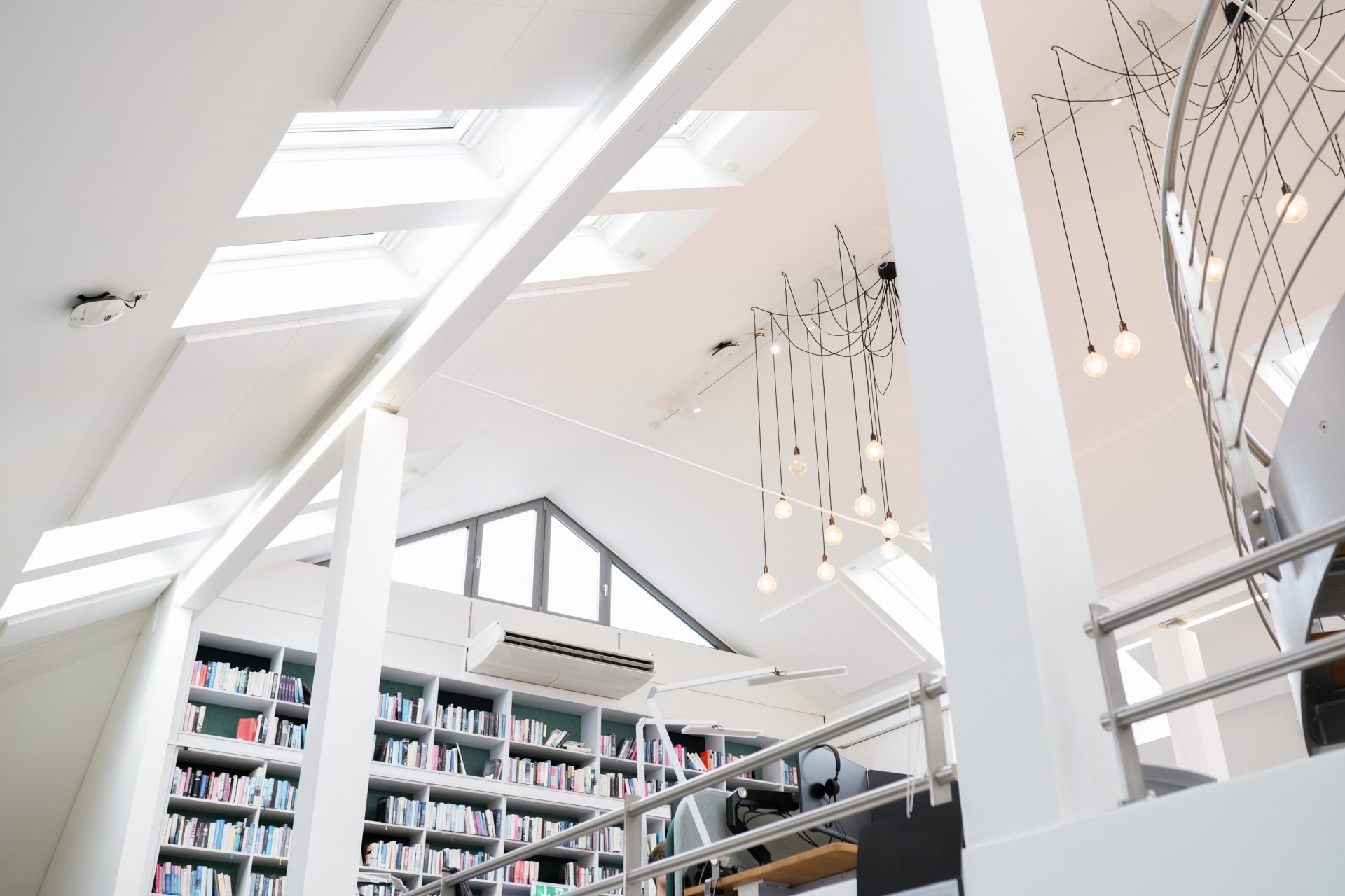 Das Bild zeigt den Innenraum einer modernen Bibliothek oder Buchhandlung. Die Architektur ist hell und offen gestaltet, mit weißen Wänden und einem hohen Dachfenster, das viel natürliches Licht hineinlässt. Auf der rechten Seite des Bildes sind große, kunstvolle Leuchten zu sehen, die aus vielen kleinen Glühbirnen bestehen, und rechts unten sind Regale mit einer Vielzahl von Büchern erkennbar. Die Gesamteindruck vermittelt eine harmonische Atmosphäre, die sowohl zum Lesen als auch zum Stöbern einlädt.