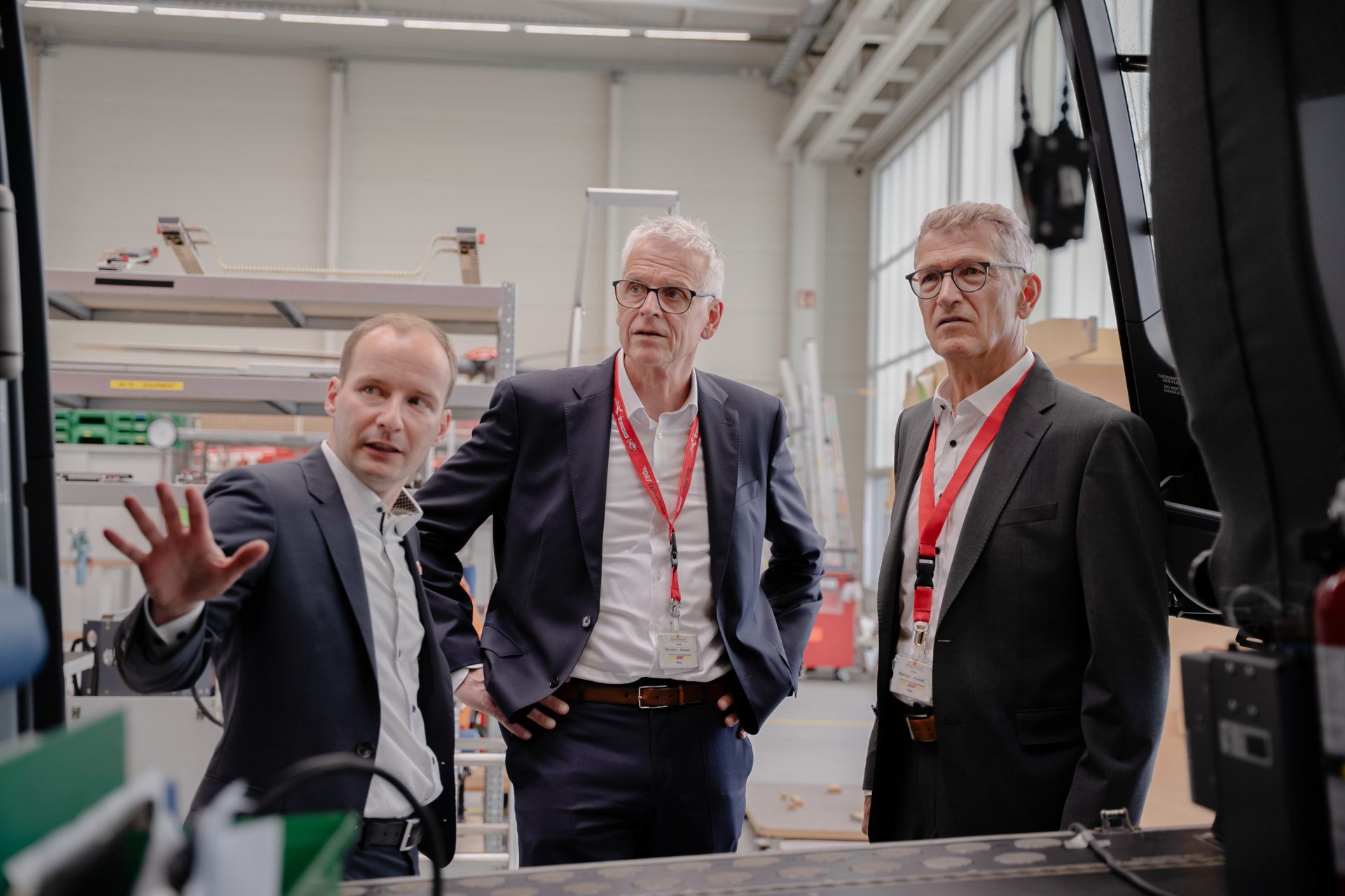 Das Bild zeigt eine Gruppe von Personen in einem geschäftlichen Umfeld, möglicherweise während einer Besichtigung oder Präsentation. Die Personen tragen Business-Kleidung und haben Umhängebänder mit Befestigungskarten, was auf eine Veranstaltung oder Konferenz hinweisen könnte. Der Raum könnte ein industrieller oder technischer Bereich sein, basierend auf den sichtbaren Maschinen und Gerätschaften.