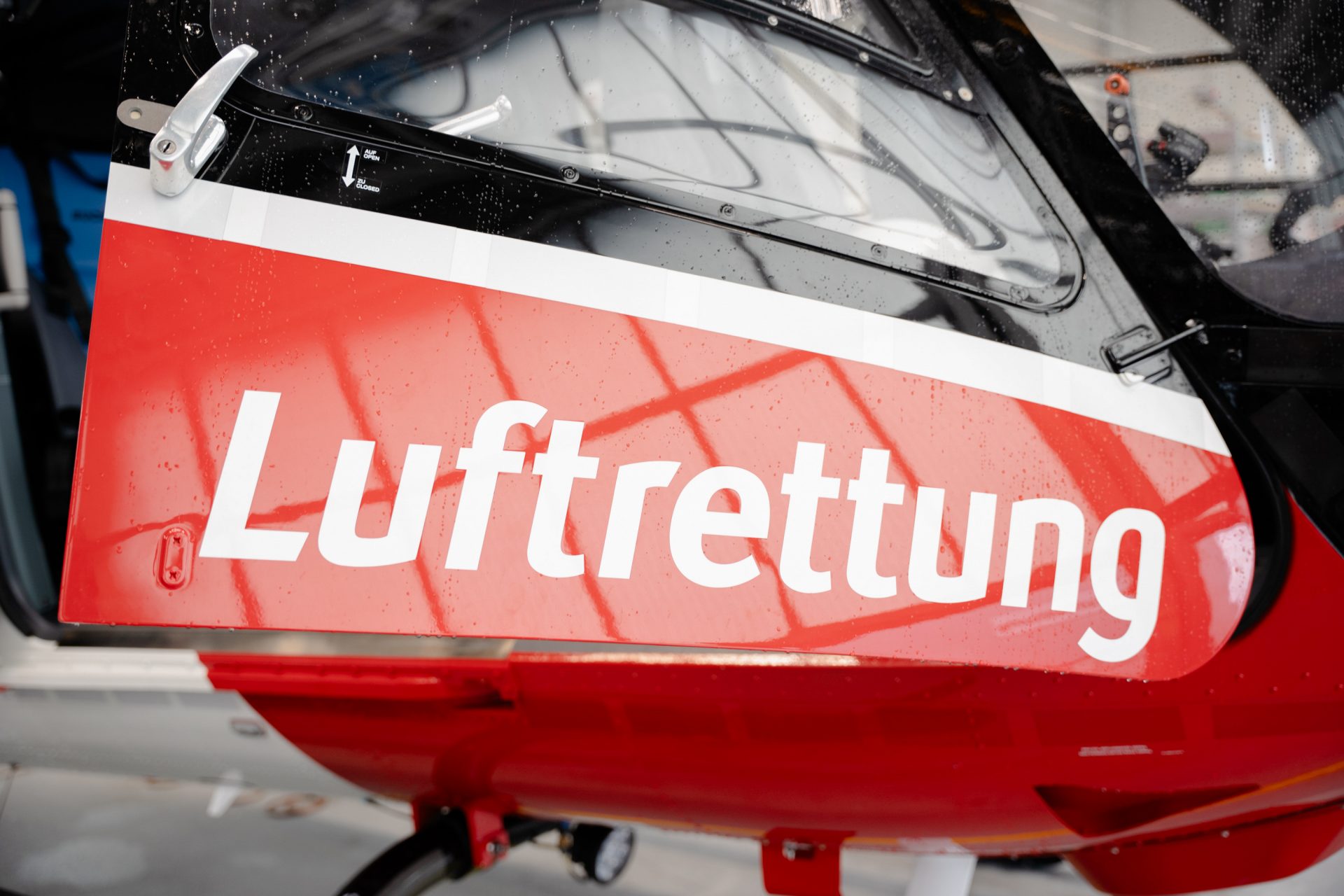 Das Bild zeigt ein Detail eines Rettungshubschraubers, genauer gesagt die Seite oder die Tür des Cockpits, auf dem mit großen Buchstaben das Wort "Luftrettung" geschrieben steht. Dies deutet darauf hin, dass der Hubschrauber Teil einer Luftrettungsorganisation ist, die sich darauf spezialisiert, schnelle medizinische Hilfe durch Lufttransport anzubieten. Die Farbgestaltung ist typisch für Rettungsfahrzeuge und signalisert deutlich deren Zweck und Funktion für Notfall- und Rettungseinsätze.