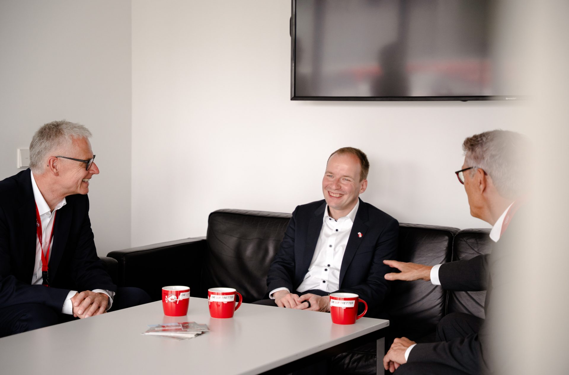 Das Bild zeigt eine Gruppe von Personen, die in einem Besprechungsraum oder Lounge-Bereich sitzen. Sie sind in einem Gespräch vertieft. Auf dem Tisch vor ihnen stehen drei rote Tassen und eine Broschüre, die möglicherweise Informationsmaterialien darstellen könnten. Im Hintergrund ist ein Fernseher zu sehen.