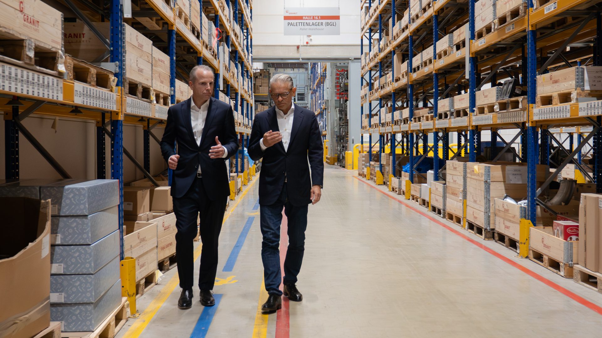 Das Bild zeigt zwei Männer mit Anzügen beim Gehen durch einen industriellen Lagerbereich. Es scheint sich um eine Geschäftsumgebung zu handeln, möglicherweise ein Lagerhaus oder eine Großhandelslage, was durch die Regale voller Kisten und Kartons suggeriert wird. Die Männer sind in ein Gespräch vertieft und scheinen berufliche Themen zu diskutieren, während sie durch den gut organisierten Raum gehen. Die Halle ist geräumig und durch Markierungen auf dem Boden strukturiert.