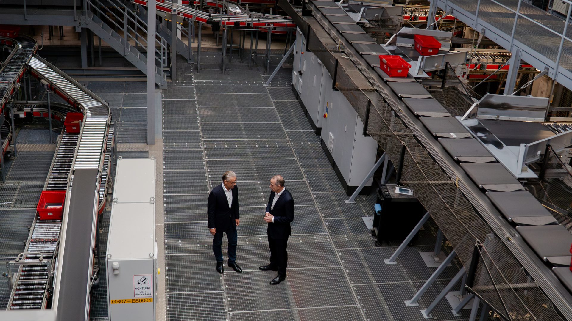 Das Bild zeigt das Innere einer industriellen Verpackungs- oder Sortieranlage. Auf dem Foto sind zwei Personen zu sehen, die miteinander sprechen, vermutlich Arbeiter oder Führungskräfte des Betriebs. Um sie herum befinden sich zahlreiche Maschinen, Förderbänder und rote Behälter, die bezeugen, dass hier Produkte weiterverarbeitet oder sortiert werden könnten. Die strukturierten Böden und technologisch ausgestatteten Bereiche lassen auf eine moderne Einrichtung schließen, die möglicherweise in einem Logistikzentrum oder einer Fabrik zu beobachten ist.
