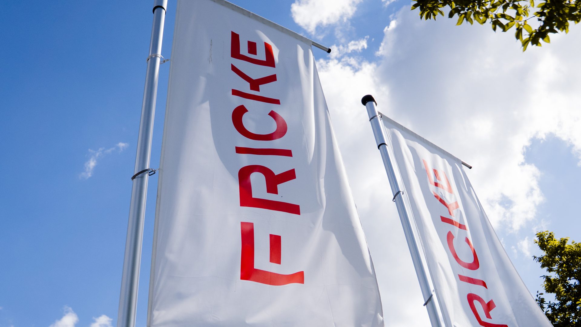 Das Bild zeigt zwei Fahnen mit dem Firmenlogo „FRICKE“, die an Fahnenmasten im Freien montiert sind. Die Fahnen sind weiß und haben den roten Schriftzug „FRICKE“, der zentral darauf abgebildet ist. Im Hintergrund sind der blaue Himmel und einige Wolken sichtbar, was eine sonnige Atmosphäre suggeriert.