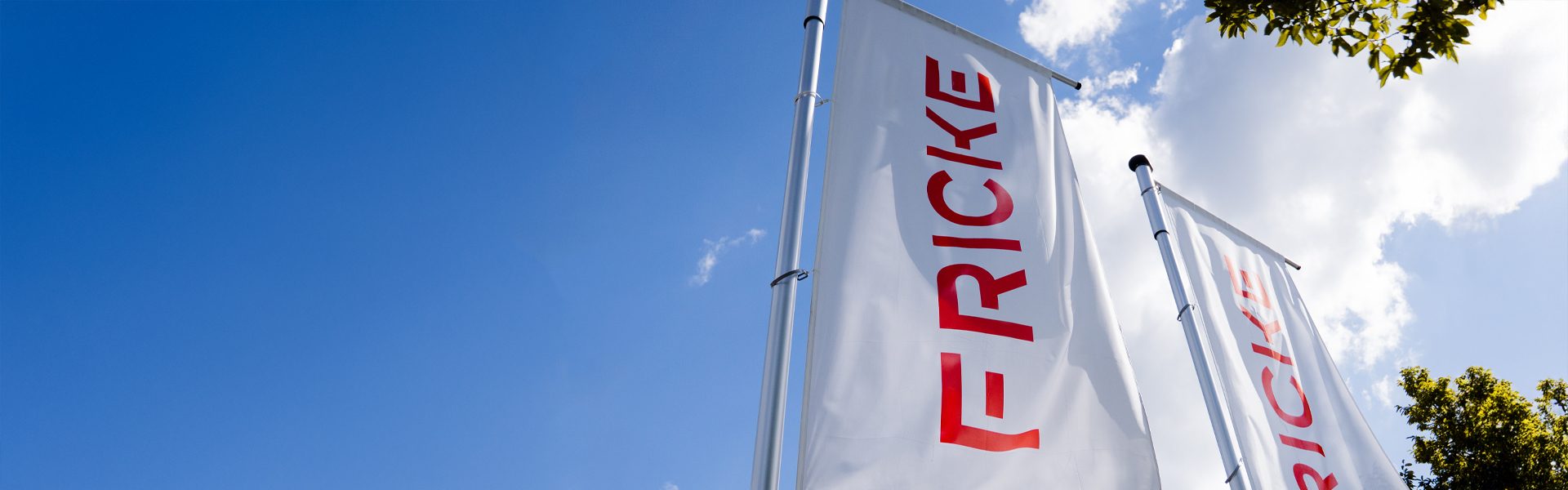 Das Bild zeigt Flaggen mit der Aufschrift "FRICKE" in einem blauen Himmel mit einigen Wolken und einem Baum im Hintergrund. Der Fokus liegt auf den Flaggen, die anscheinend zu einem Unternehmen oder einer Marke gehören. Es gibt keinen weiteren Kontext oder Details zur Umgebung im Bild.