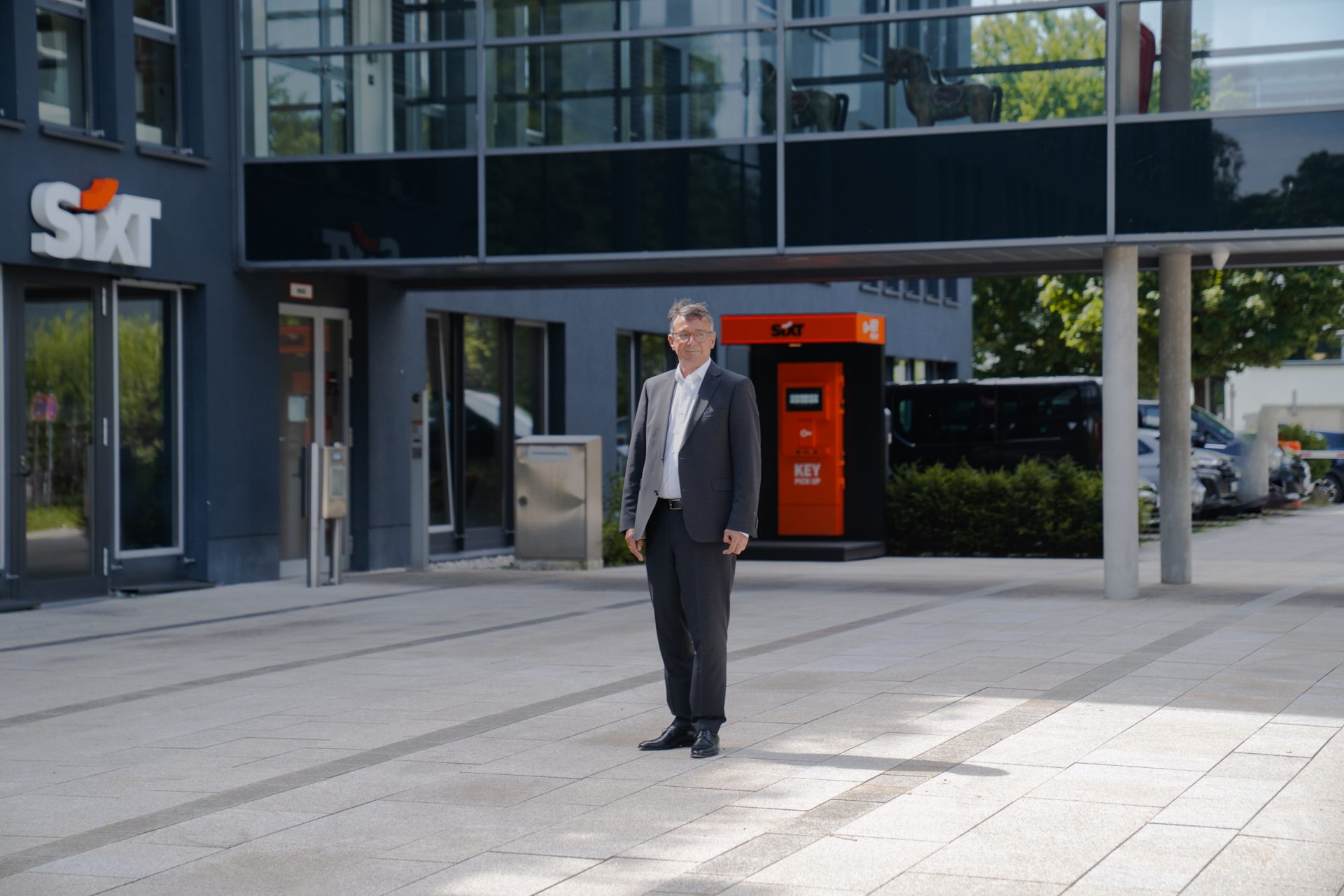 Das Bild zeigt einen Mann im Anzug, der vor einem Gebäude steht. Auf der linken Seite ist das Logo von Sixt, einem bekannten Autovermieterunternehmen, sichtbar. Das Gebäude scheint eine moderne Geschäftsarchitektur zu haben, mit Glasfassaden und einem gepflegten Vorhof. Der Mann ist leicht unscharf, möglicherweise der Fokus des Fotografen, während das Gebäude und das Sixt-Logo klarer erkennbar sind. Im Hintergrund ist ebenfalls ein roter Automat oder ein Automatengerät zu sehen, was auf einen modernen Unternehmenssitz oder eine Filiale hinweist.