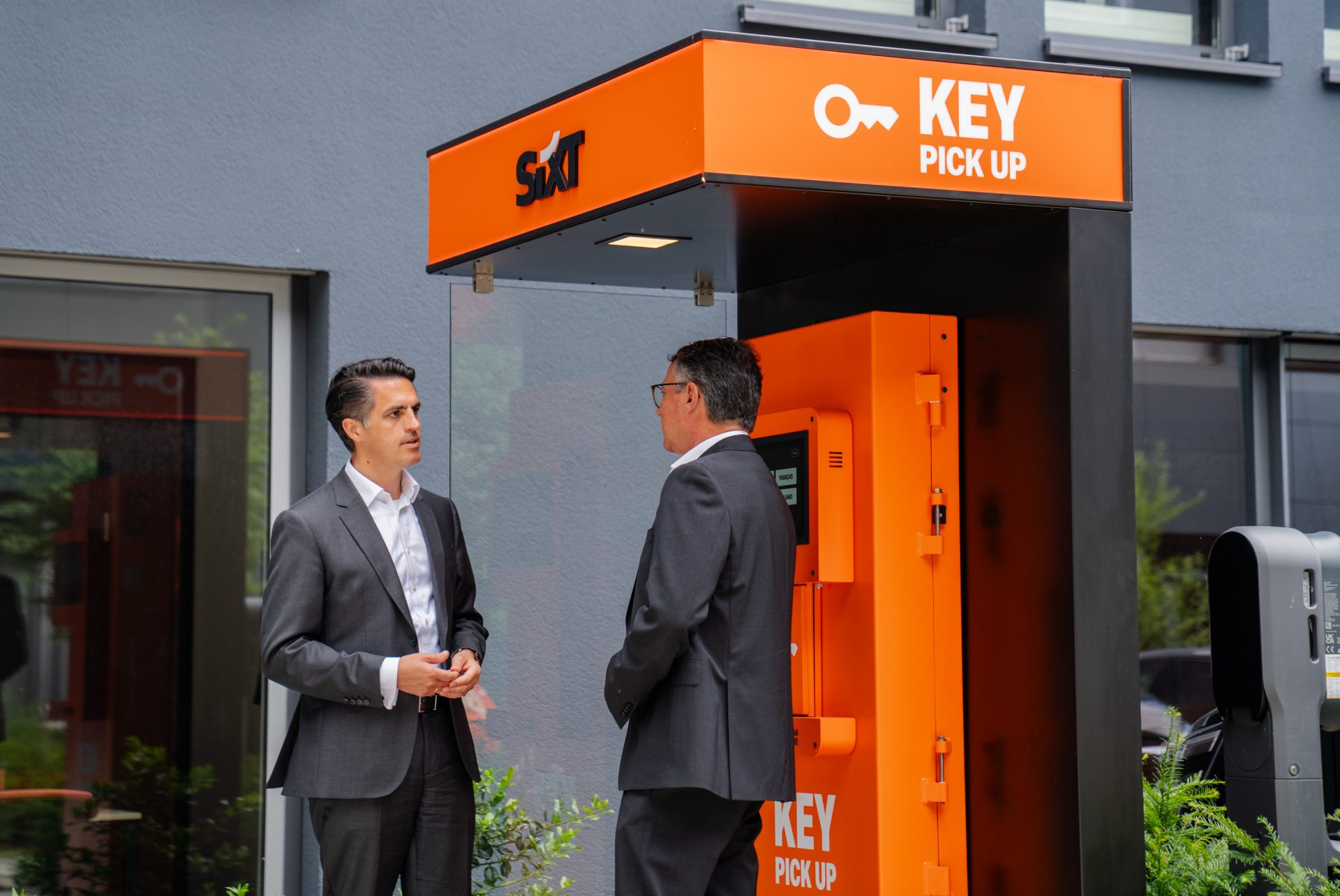 Das Bild zeigt zwei Männer in formeller Kleidung, die vor einem orangenen Key Pick-Up Kiosk stehen. Der Kiosk trägt das Logo von "Sixt", einem bekannten deutschen Mietwagenanbieter. Der Kiosk scheint Teil eines automatisierten Systems zur Schlüsselübergabe für Mietfahrzeuge zu sein, was es den Kunden ermöglicht, ihre Fahrzeugschlüssel einfach und schnell abzuholen, vermutlich ohne direkte Interaktion mit einem Mietwagenangestellten. 

Der Standort dieses Kiosks scheint in der Nähe eines Gebäudes oder Büros zu sein, möglicherweise am Eingang einer Sixt Mietwagenstation oder eines Partner-Unternehmens. Durch das Design und die Funktionalität des Kiosks wird ein moderner und effizienter Service vermittelt.