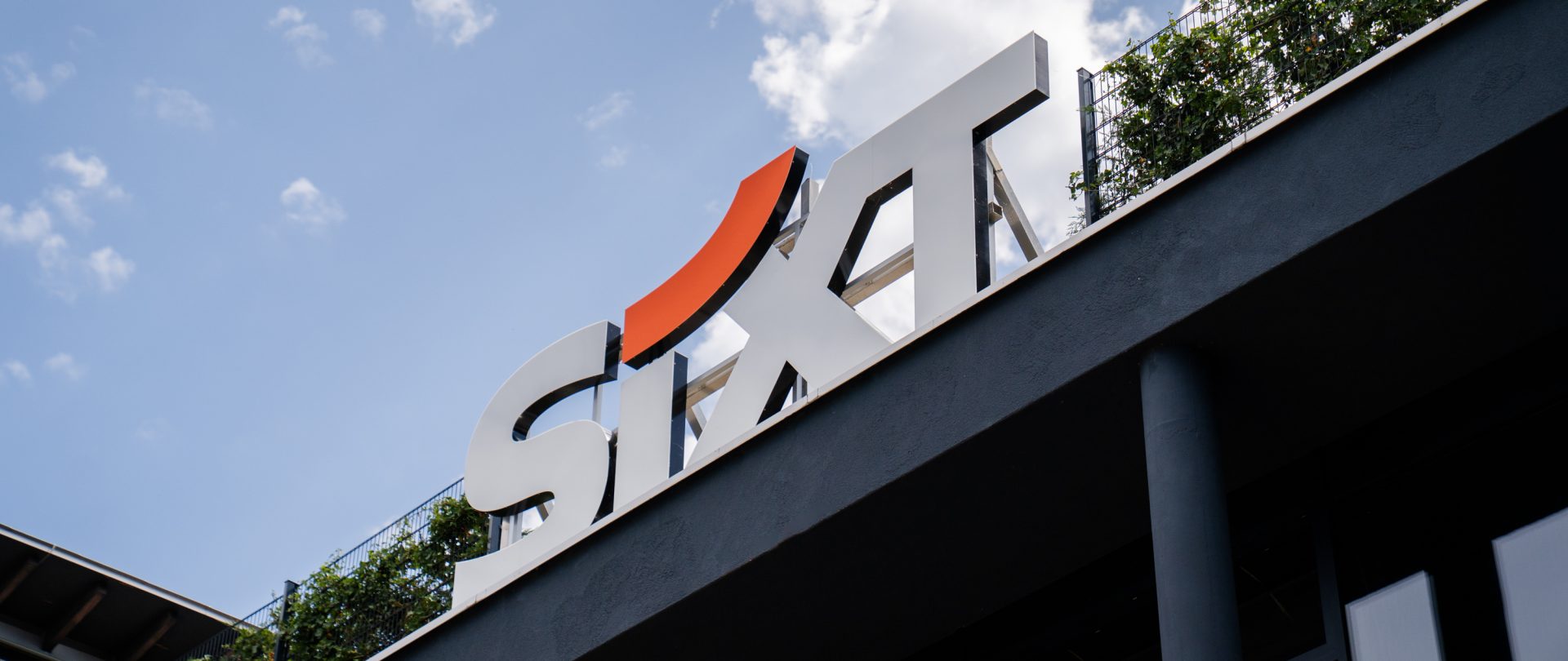 Das Bild zeigt ein Gebäude mit dem Logo der Firma SIXT auf einem Schild. SIXT ist ein berühmtes internationales Unternehmen für Autovermietung und Mobilitätsdienstleistungen. Das Logo ist stilisiert mit einem markanten orangefarbenen Element, das einem kleinen Bogen ähnelt und in das "S" integriert ist. Die Aufnahme der Marke ist gut sichtbar und im Vordergrund, wodurch der Fokus auf das Unternehmenszeichen gelegt wird. Das Bild könnte von einer Niederlassung oder Geschäftsstelle der Firma stammen.