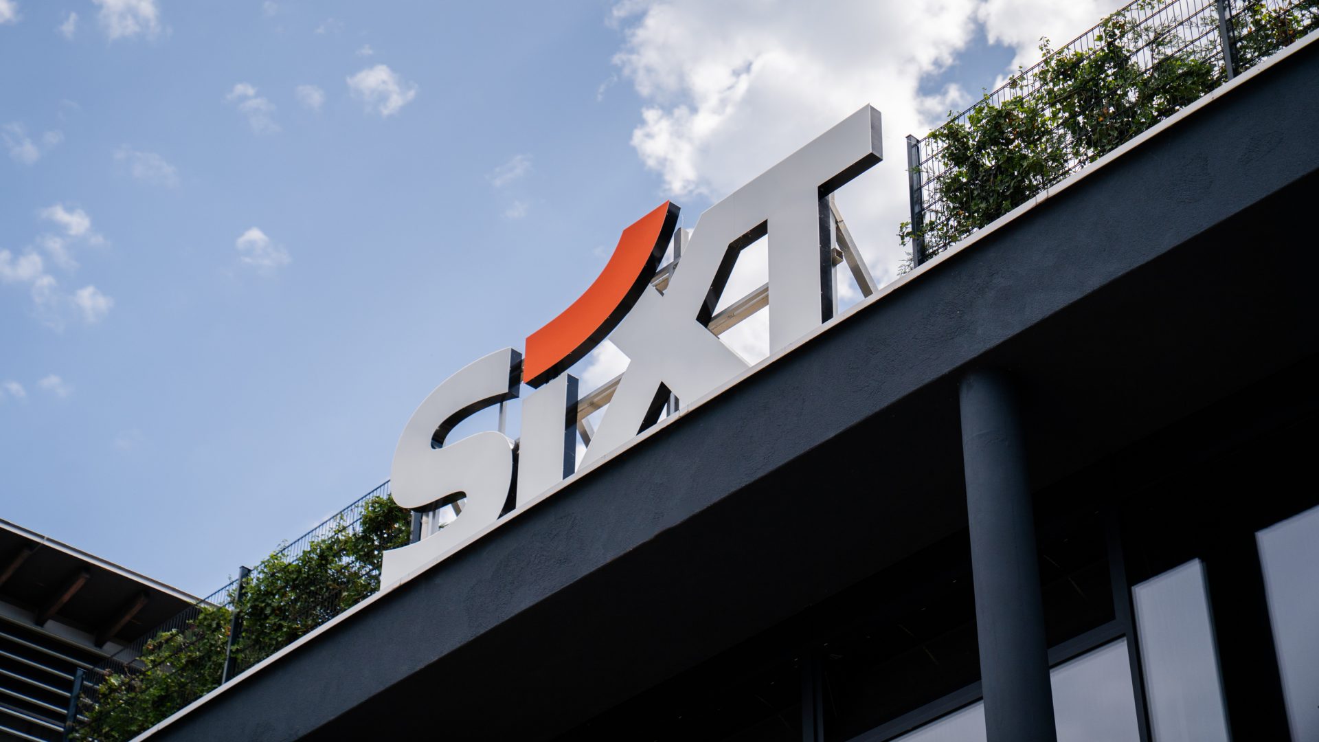 Das Bild zeigt ein Gebäude mit dem Logo der Firma SIXT, einem internationalen Unternehmen, das sich auf die Vermietung und das Leasing von Fahrzeugen spezialisiert hat. Das Logo enthält den Firmennamen "SIXT" in weiß mit einem markanten orangefarbenen Akzent über dem Buchstaben "I". Das Gebäude scheint Teil der Infrastruktur des Unternehmens zu sein, möglicherweise eines seiner Büros oder eine seiner Filialen. Über dem Logo und dem Gebäude sind Teile des blauen Himmels und einige Wolken sichtbar, was einen offenen und einladenden Eindruck vermittelt.