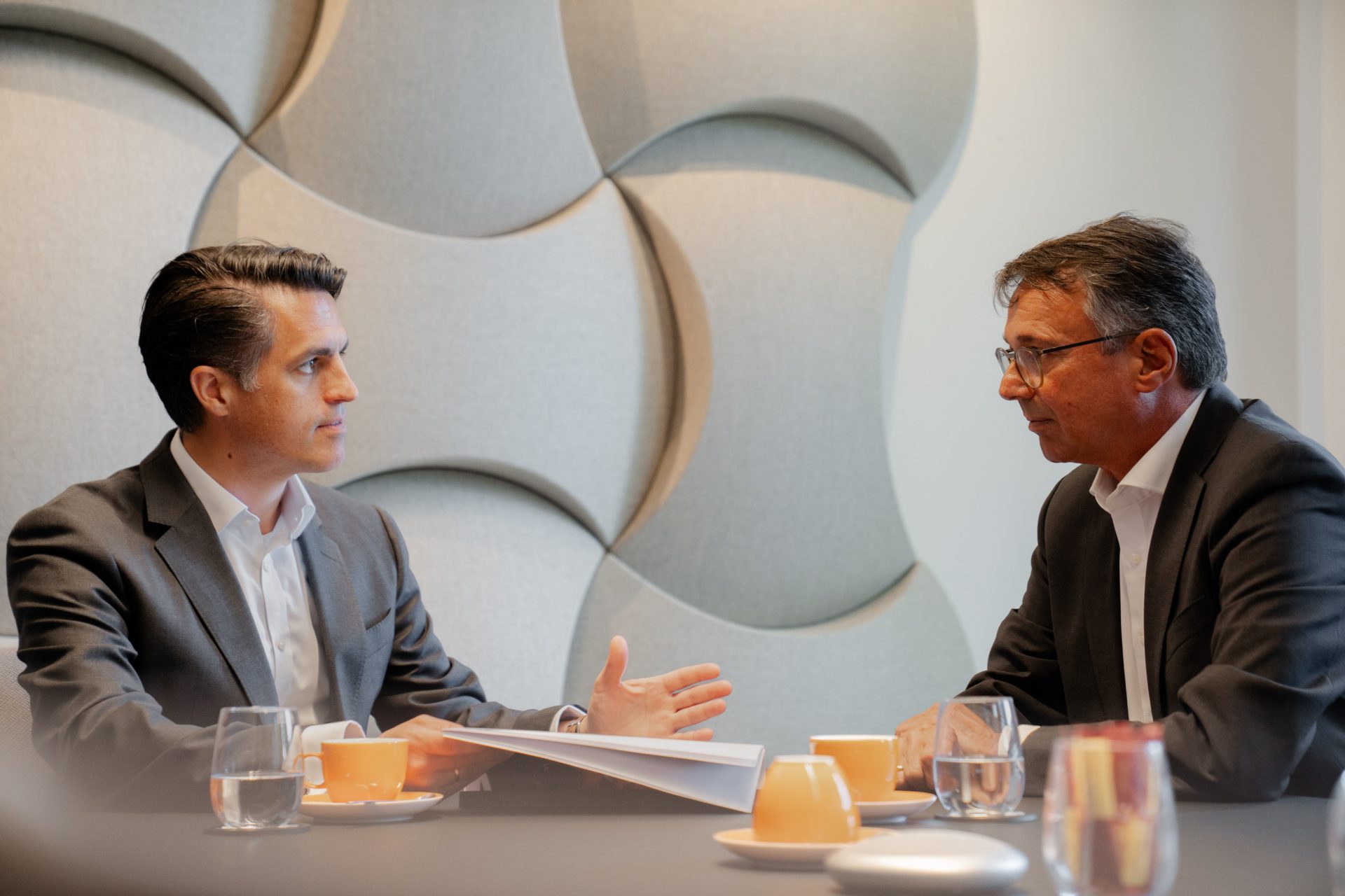 Das Bild zeigt eine Umgebung, die aussieht wie ein geschäftliches Treffen oder eine Besprechung. Zwei Personen sitzen sich gegenüber an einem Tisch. Vor ihnen liegen einige Gegenstände, darunter Gläser, Kaffeetassen, und ein Notizbuch. Im Hintergrund sieht man eine Wand mit geometrischen Formen als Dekoration.