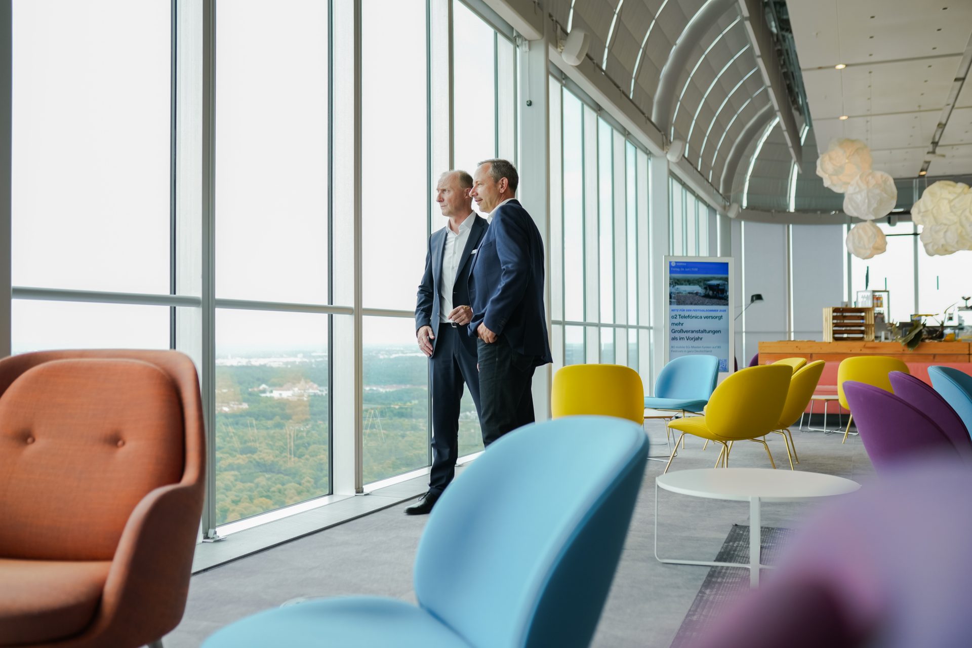 Das Bild zeigt ein modernes Büro oder eine Lounge mit zwei männlichen Personen, die in Business-Anzüge gekleidet sind und an einem großen Fenster mit Blick auf die Stadtlandschaft stehen. Der Raum ist mit stilvollen, farblich gemischten Designer-Stühlen und einem großen Tisch ausgestattet, was auf eine zeitgenössische und offene Arbeitsumgebung hinweist. Die Architektur des Raumes wirkt minimalistisch und hell, was die Bedeutung von Offenheit und Kreativität in modernen Geschäftsräumen hervorhebt.