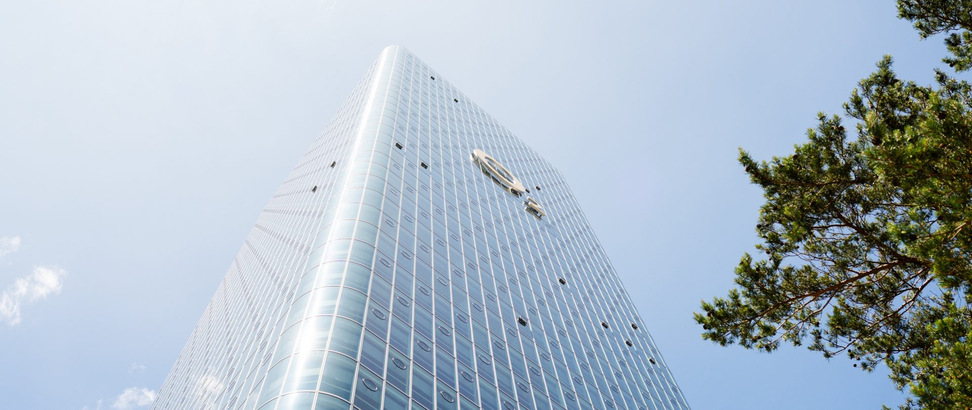Das Bild zeigt ein modernes Hochhaus, das sich gegen den Himmel erstreckt. Der Bau hat eine auffällige Glasfassade, die mit reflektierenden Paneelen bedeckt ist, sodass der Himmel und die Umgebung auf der Oberfläche sichtbar sind. Auf dem Gebäude sind Fenster oder Strukturen zu erkennen, die gleichmäßig über die gesamte Höhe verteilt sind, was auf ein funktionales und ästhetisches Design hindeutet. Rechts im Bild ist ein Baum zu sehen, der einen natürlichen Kontrast zu der städtischen Architektur bildet.