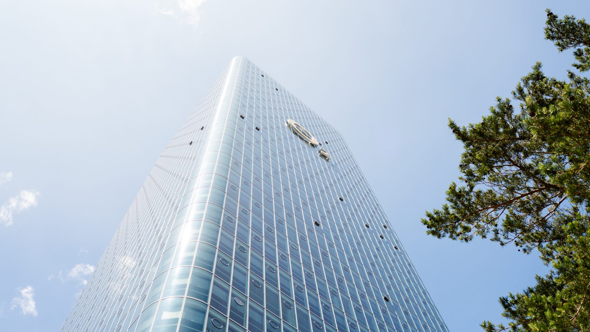 Das Bild zeigt ein modernes Hochhaus, fotografiert aus einer Perspektive von unten nach oben, die die beeindruckende Höhe und Architektur des Gebäudes betont. Das Hochhaus hat eine Glasfassade, die das Licht reflektiert und die klare Struktur des Gebäudes hervorhebt. Das Glas bietet einen Blick auf den Himmel, der teilweise von Wolken durchzogen ist, und einen Baum, der am rechten Bildrand sichtbar ist, was einen Kontrast zwischen der urbanen und der natürlichen Umgebung erzeugt. Die Perspektive vermittelt ein Gefühl der Erhabenheit und Größe dieses Bauwerks.