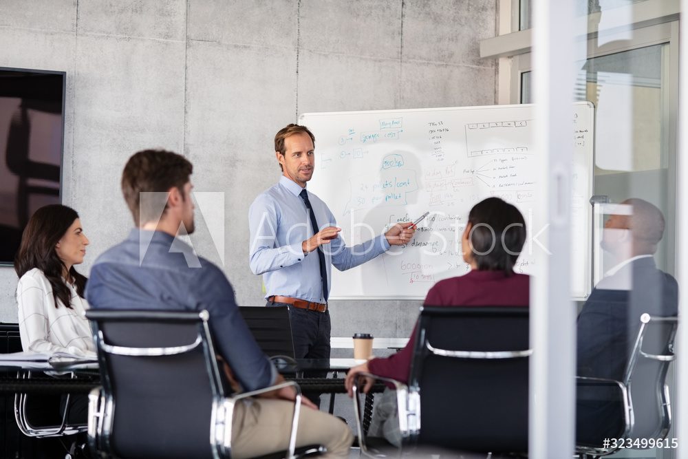 Das Bild zeigt eine Gruppe von Menschen, die an einer geschäftlichen Besprechung teilnehmen. Im Zentrum steht ein Mann, der anscheinend mit einer Präsentation oder Erklärung beschäftigt ist, während er auf ein Whiteboard zeigt. Das Whiteboard ist mit Skizzen und Notizen gefüllt, die möglicherweise Strategien, Projekte oder Ziele der Firma darstellen. Um ihn herum sitzen mehrere Kollegen in einem modernen Konferenzraum.

Darüber hinaus scheint diese Szene in einem professionellen Geschäftsumfeld statt zu finden, um eine ernsthafte Diskussionsrunde oder eine formelle Präsentation zu symbolisieren. Die Körperhaltung und Ausdrücke der Beteiligten deuten darauf hin, dass dies ein konzentriertes und produktives Meeting ist.