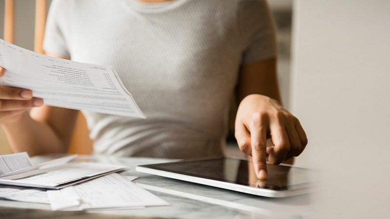 Das Bild zeigt eine Person, wahrscheinlich eine Frau, die an einem Tisch sitzt. Sie hält einen Papierstapel oder mehrere Dokumente in einer Hand und benutzt mit der anderen Hand einen Tablet-PC oder ein Smart-Gerät, das auf dem Tisch liegt. Die Szene vermittelt den Eindruck von Büroarbeit oder der Verwaltung von persönlichen oder finanziellen Angelegenheiten.