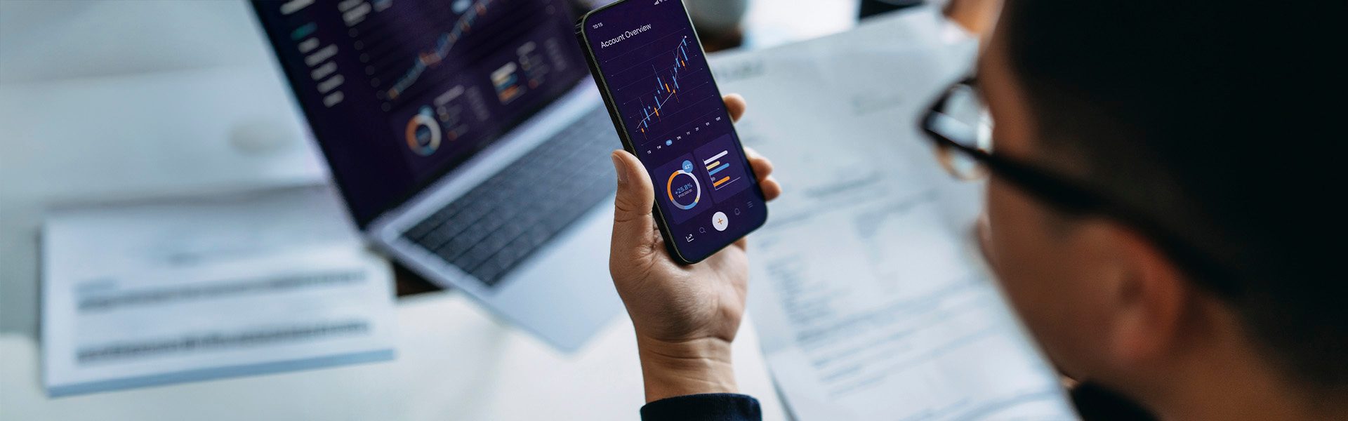 Das Bild zeigt eine Person, die ein Smartphone in der Hand hält, mit einem Laptop und Papieren im Hintergrund. Auf dem Bildschirm des Handys sind Diagramme oder Finanzdaten zu sehen, was möglicherweise auf eine Analyse oder Betrachtung von Geschäftsdaten hinweist.