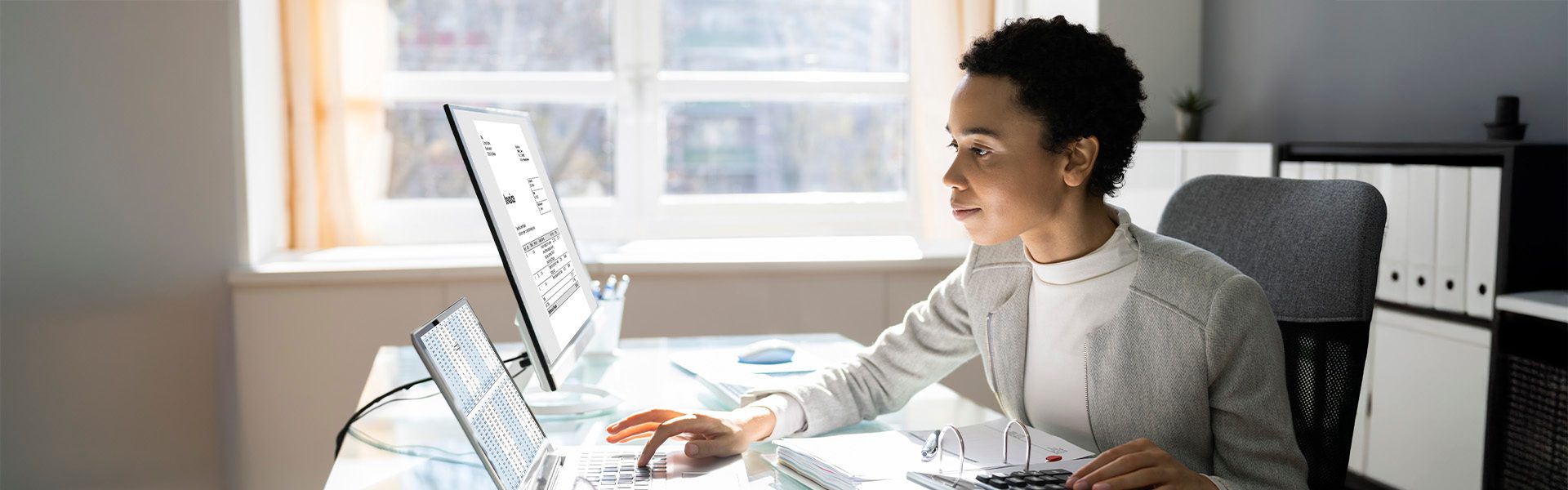 Das Bild zeigt eine Person in einem hellen Büroraum mit mehreren Monitoren, einem Laptop und Büromaterialien auf dem Tisch. Das Büro hat große Fenster, die viel Tageslicht hineinlassen. An der Rückseite sind Regale mit Ordnern zu sehen.