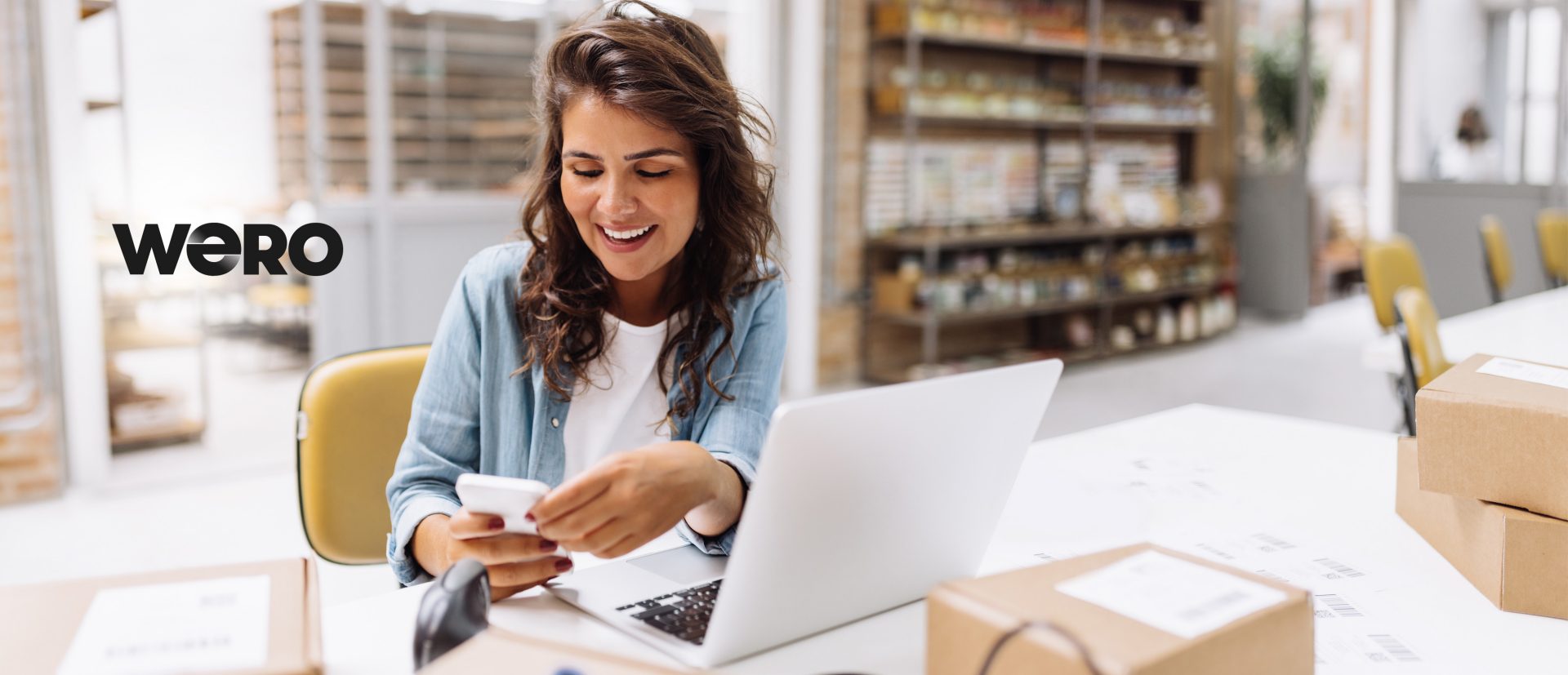 Eine Person sitzt an einem Tisch in einem Büro oder einer Lagerumgebung und benutzt ein Handy sowie einen Laptop. Es gibt Kartons auf dem Tisch, was auf eine Verpackungs- oder Versandtätigkeit hindeuten könnte. Im Hintergrund sind Regale mit verschiedenen Gegenständen oder Produkten sichtbar. Auf der linken Seite des Bildes ist das Wort "WERO" zu sehen.