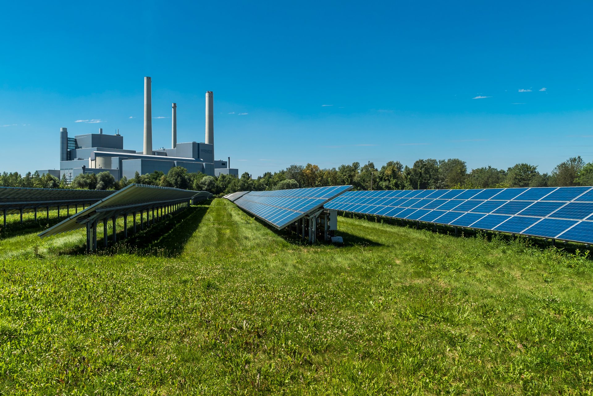 Das Bild zeigt eine Kombination aus einer Solaranlage (Photovoltaik-Anlage) im Vordergrund und einem klassischen Kraftwerk mit Schornsteinen im Hintergrund. Es verdeutlicht den Kontrast bzw. das Zusammenspiel zwischen erneuerbaren Energien und konventioneller Energieerzeugung.