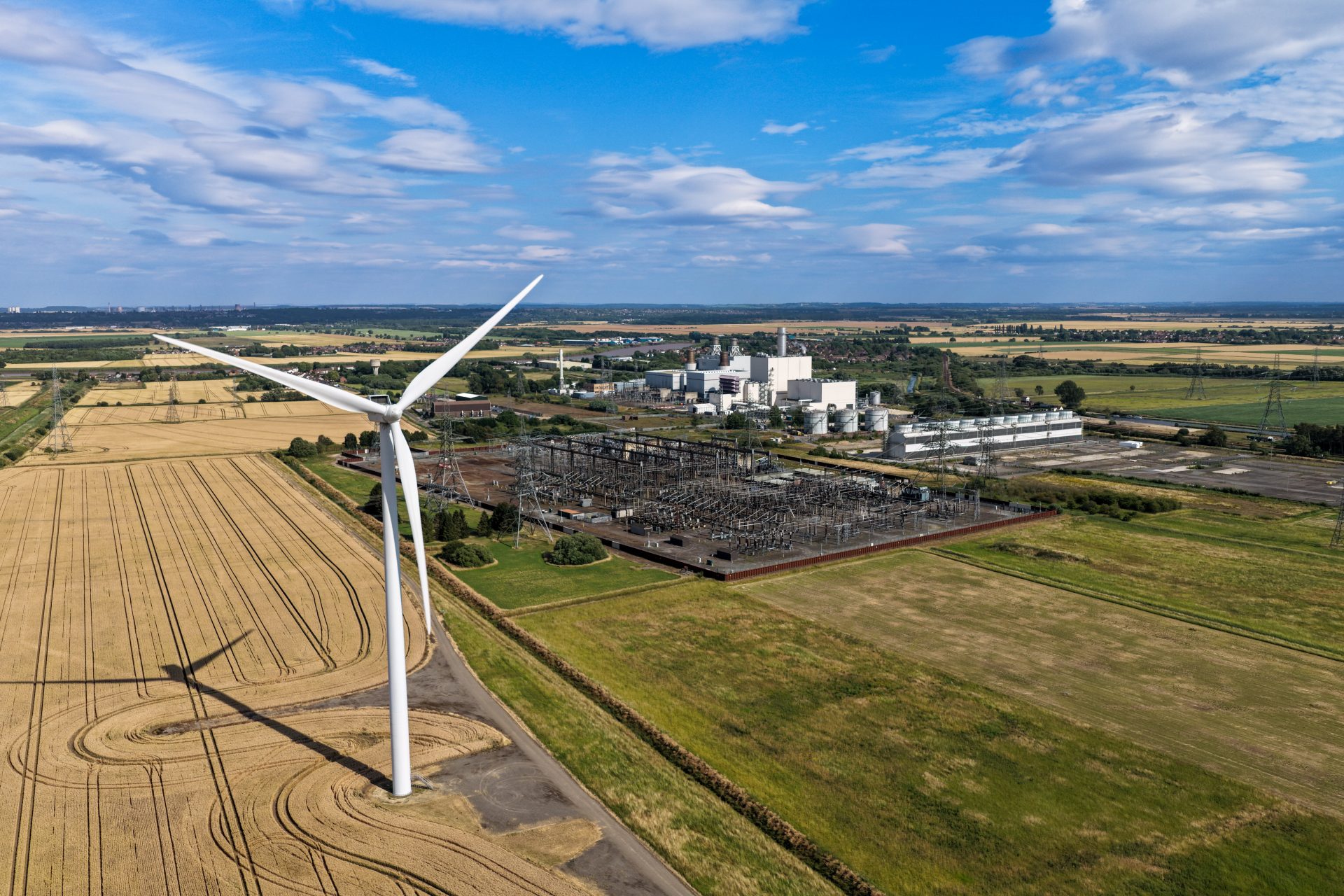 Ein Windrad vor Feldern und einem Industriekomplex veranschaulicht den Umbau des Energiesystems: Heimische erneuerbare Energien sollen fossile Energieträger zunehmend ersetzen, die Versorgungssicherheit stärken und die Abhängigkeit von Energieimporten verringern.
