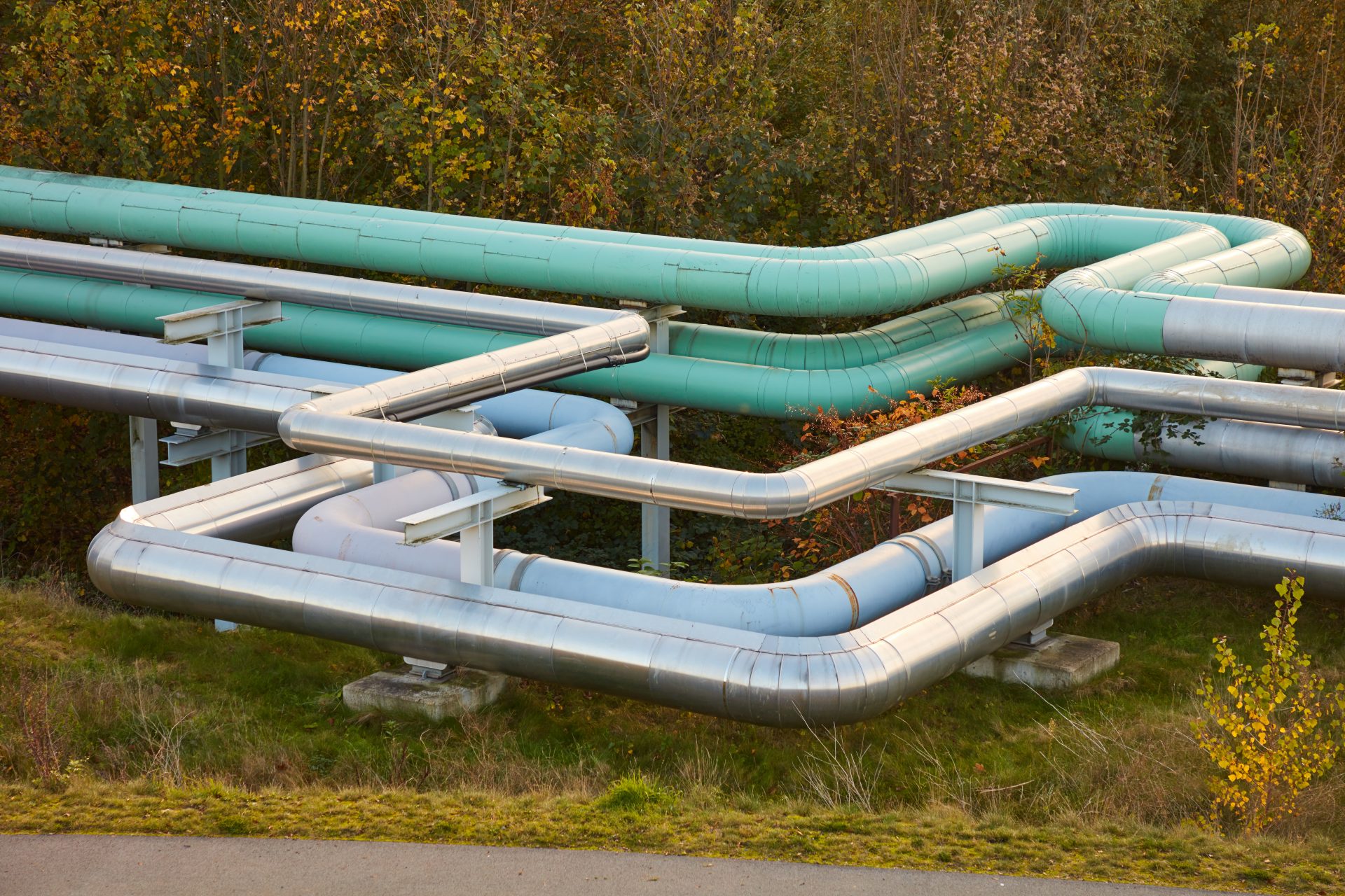 Das Bild zeigt mehrere große Rohrleitungen, die oberirdisch verlaufen. Es handelt sich vermutlich um Industrie- oder Fernwärmeleitungen, die für den Transport von Gas, Wasser oder anderen Medien verwendet werden. Die Rohre verlaufen parallel und machen mehrere Biegungen. Im Hintergrund ist Vegetation zu sehen.