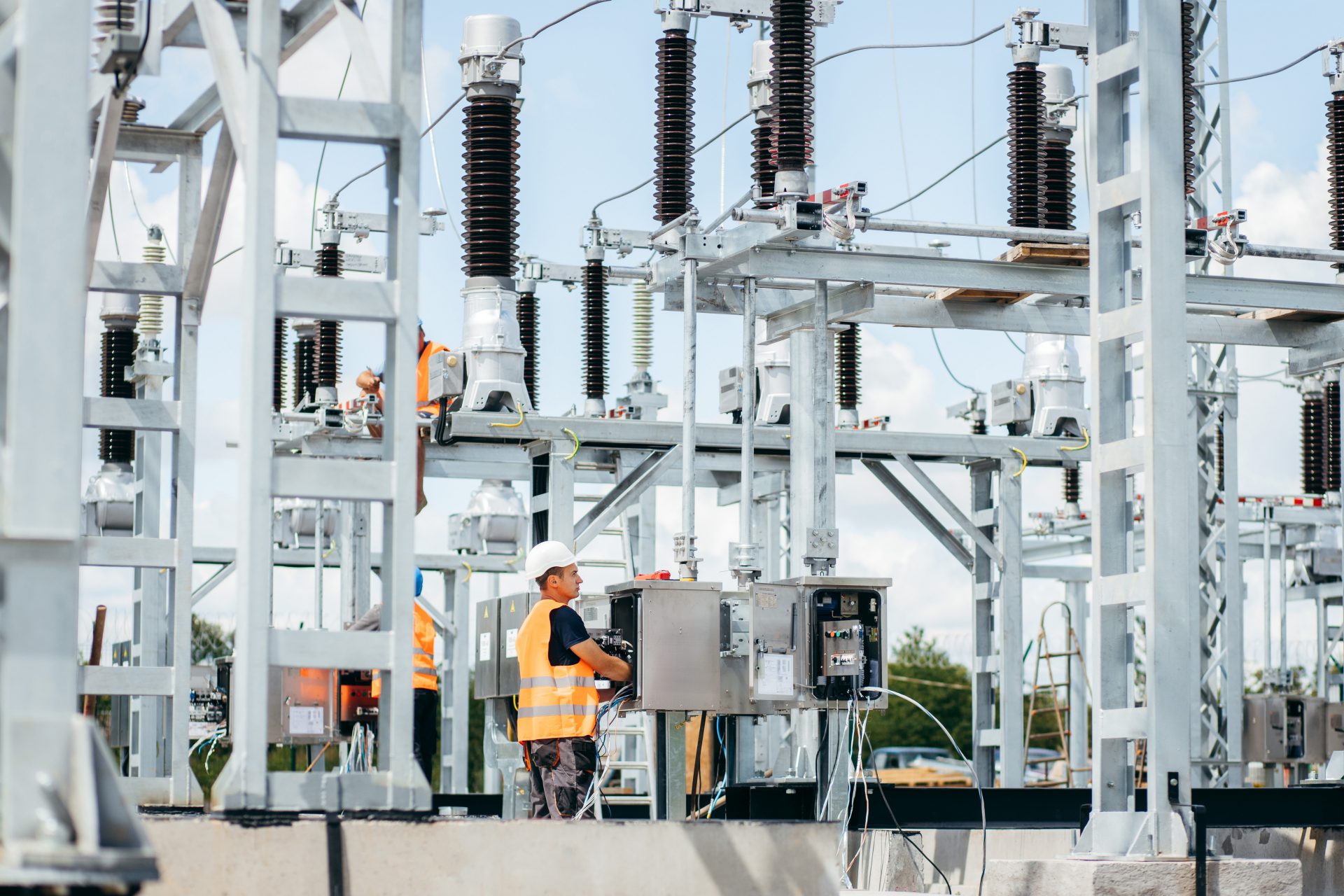 Das Bild zeigt eine elektrische Umspannanlage (Umspannwerk) mit mehreren Transformatoren und Schaltanlagen. Im Vordergrund arbeitet eine Person in Warnweste an einem Schaltschrank. Solche Anlagen werden verwendet, um elektrische Energie umzuwandeln und zu verteilen.