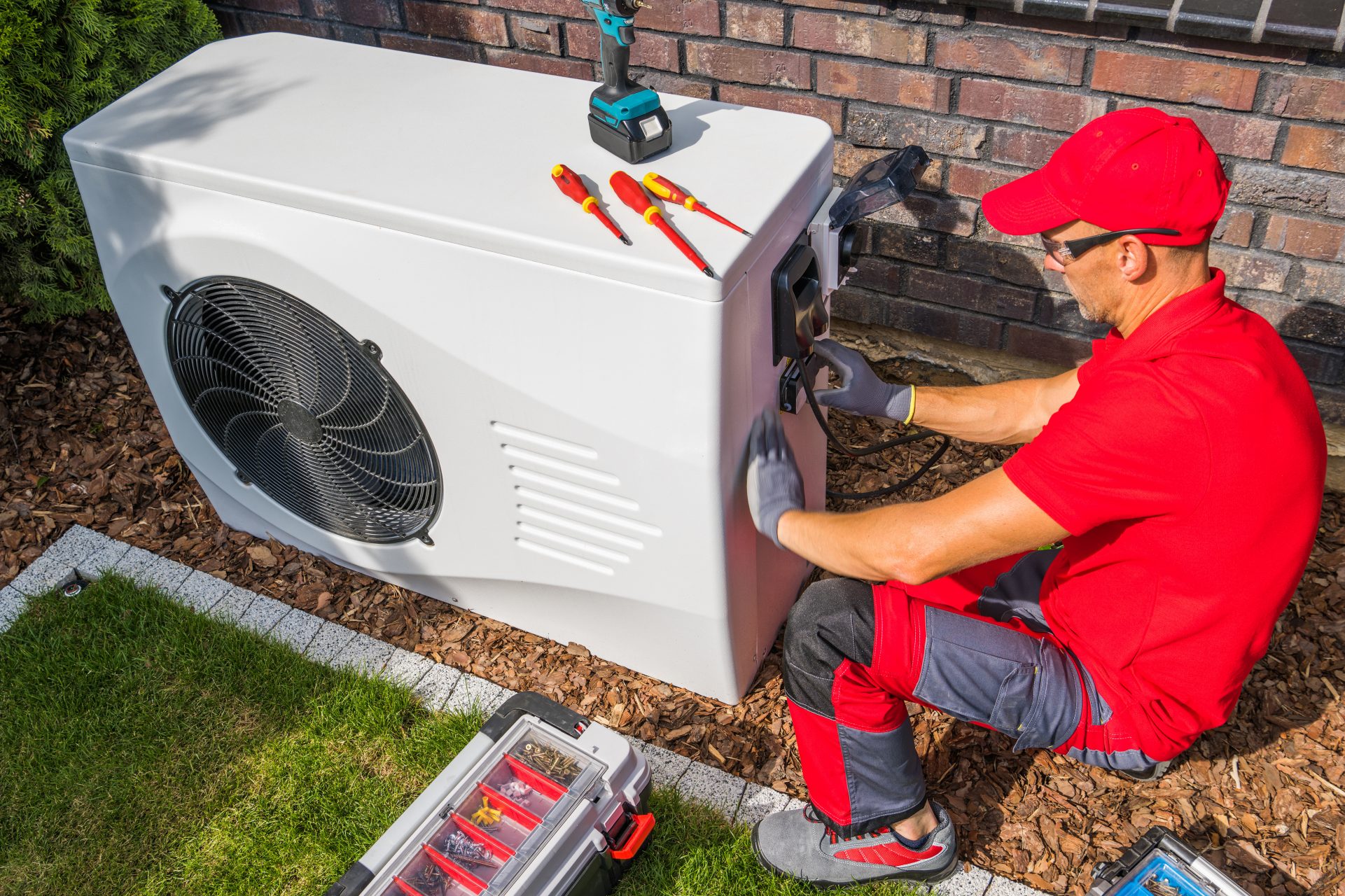 Ein Handwerker installiert oder wartet die Außeneinheit einer Wärmepumpe an einer Hauswand. Die Technik steht für die Dekarbonisierung der Gebäudewärme und den Investitionsbedarf privater und gewerblicher Immobilienbesitzer.
