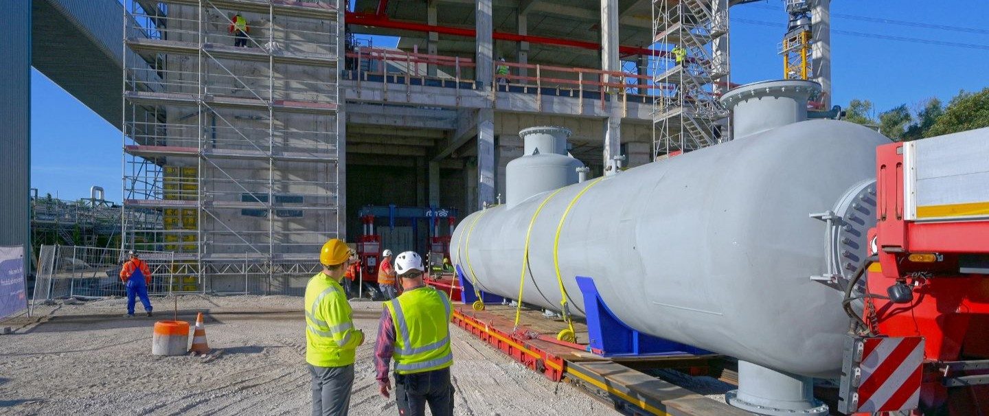 Das Bild zeigt eine Baustelle, auf der mehrere Bauarbeiter mit Schutzhelmen und Warnwesten zu sehen sind. Im Vordergrund befindet sich ein großes zylindrisches Industriebauteil oder -behälter, das vermutlich installiert oder transportiert wird. Im Hintergrund sieht man ein Gebäude, das von einem Baugerüst umgeben ist, was auf laufende Bau- oder Installationsarbeiten hindeutet.